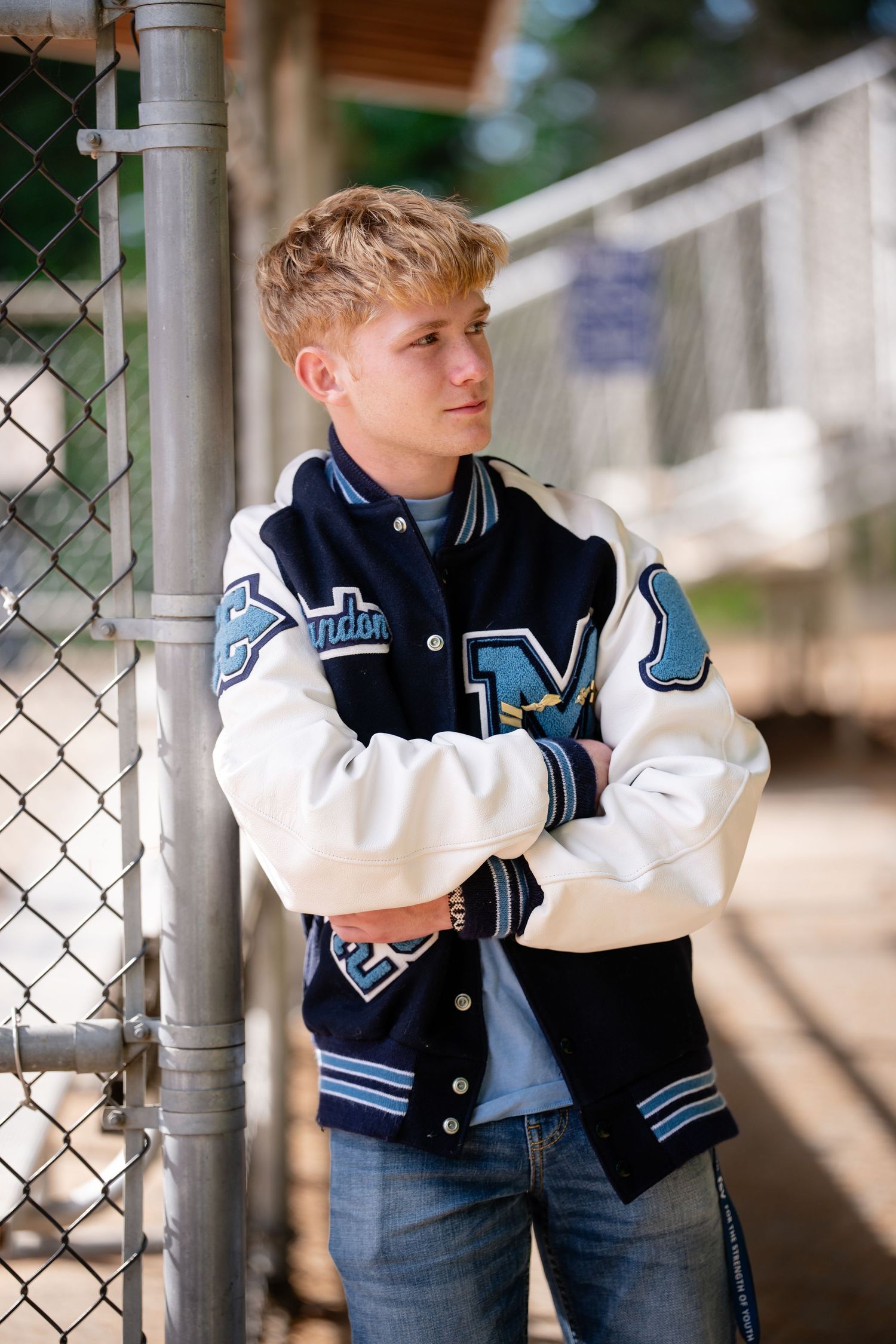 Student wearing varsity letterman jacket poses against chain link fence.