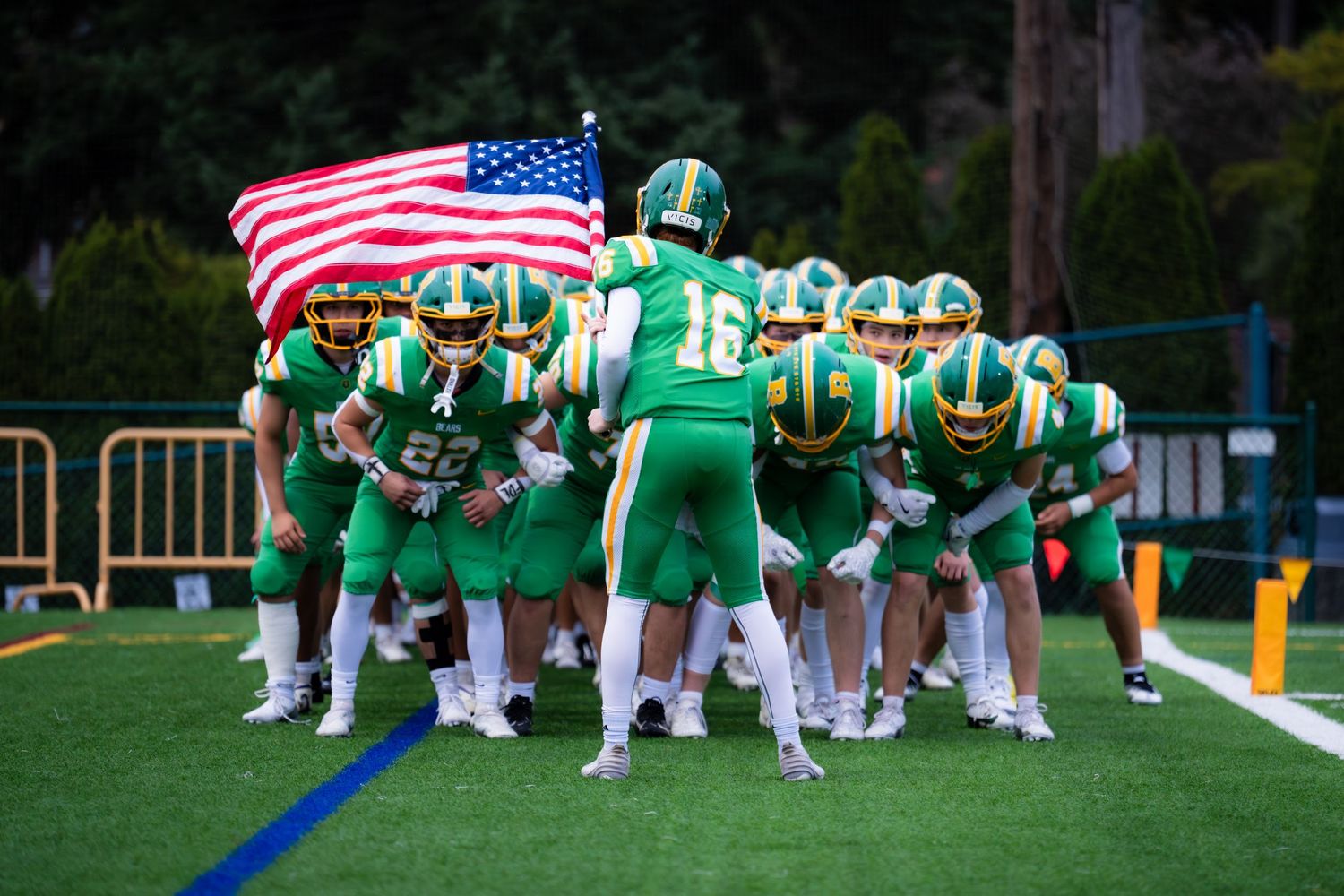 High school football team in green uniforms stands united on field while player carries American flag before game.