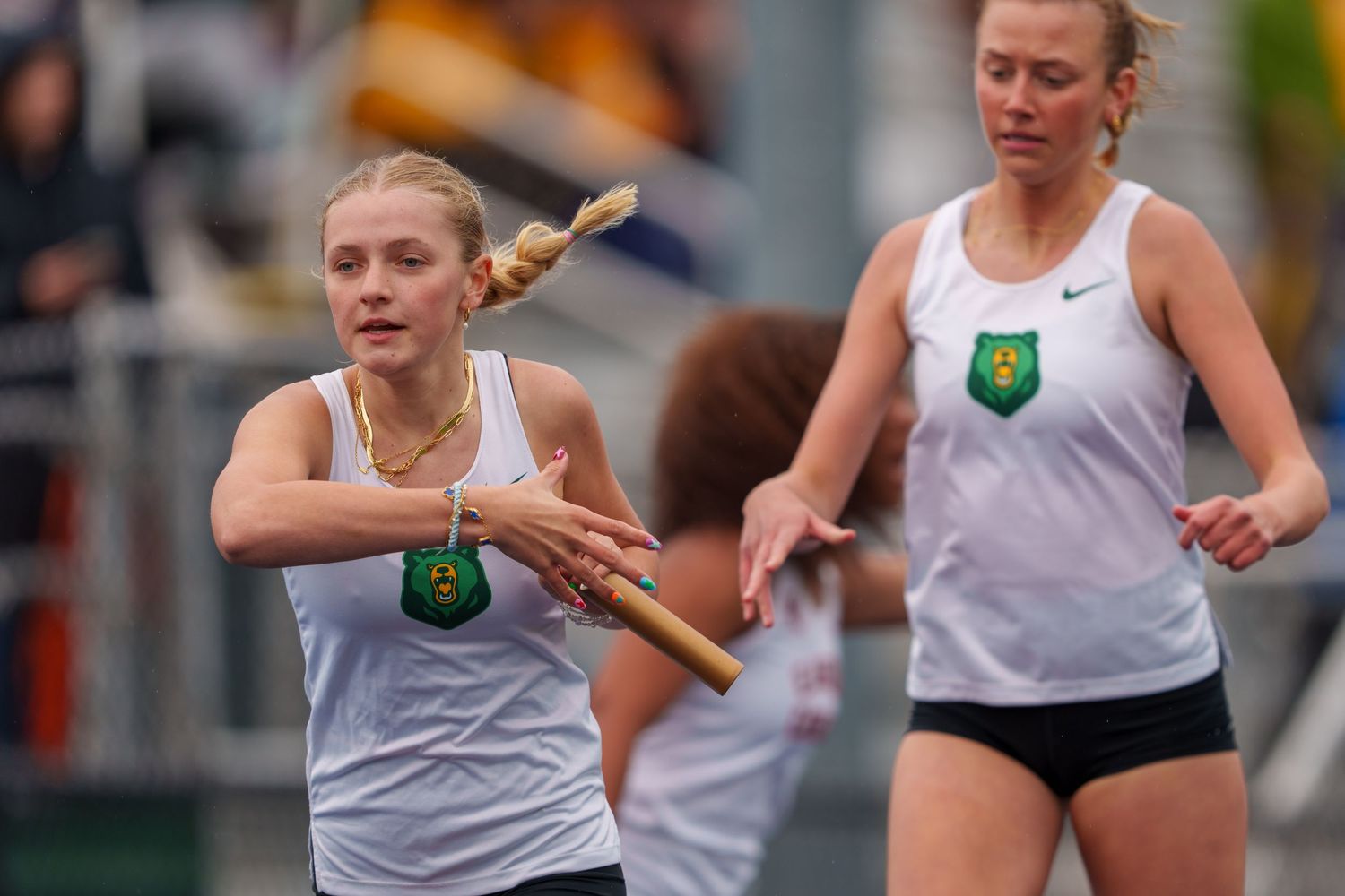 Athletes in white tank tops compete in a track relay race passing batons during an outdoor competition.
