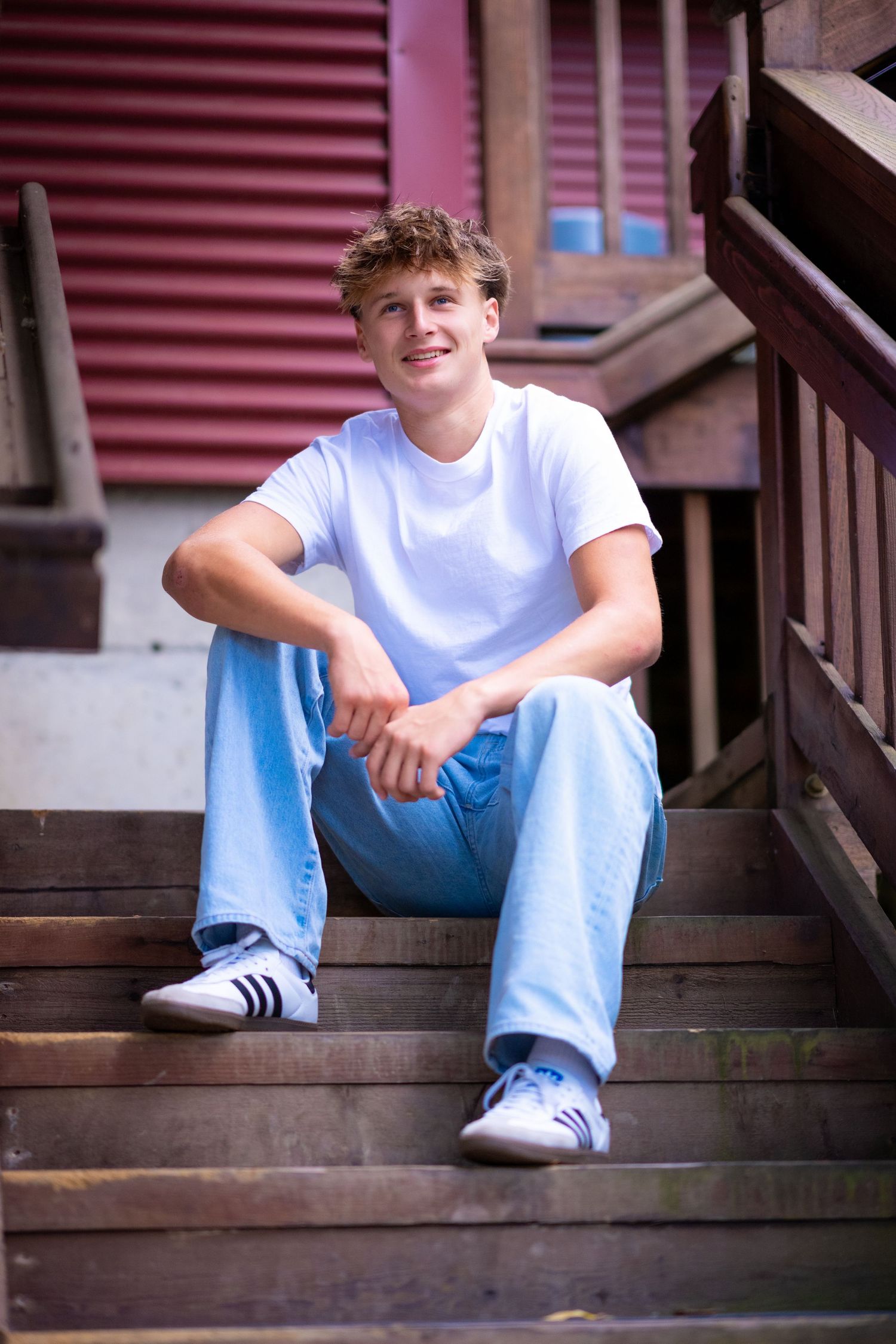 A person in a white t-shirt and light blue jeans sits casually on wooden steps outside a red building.
