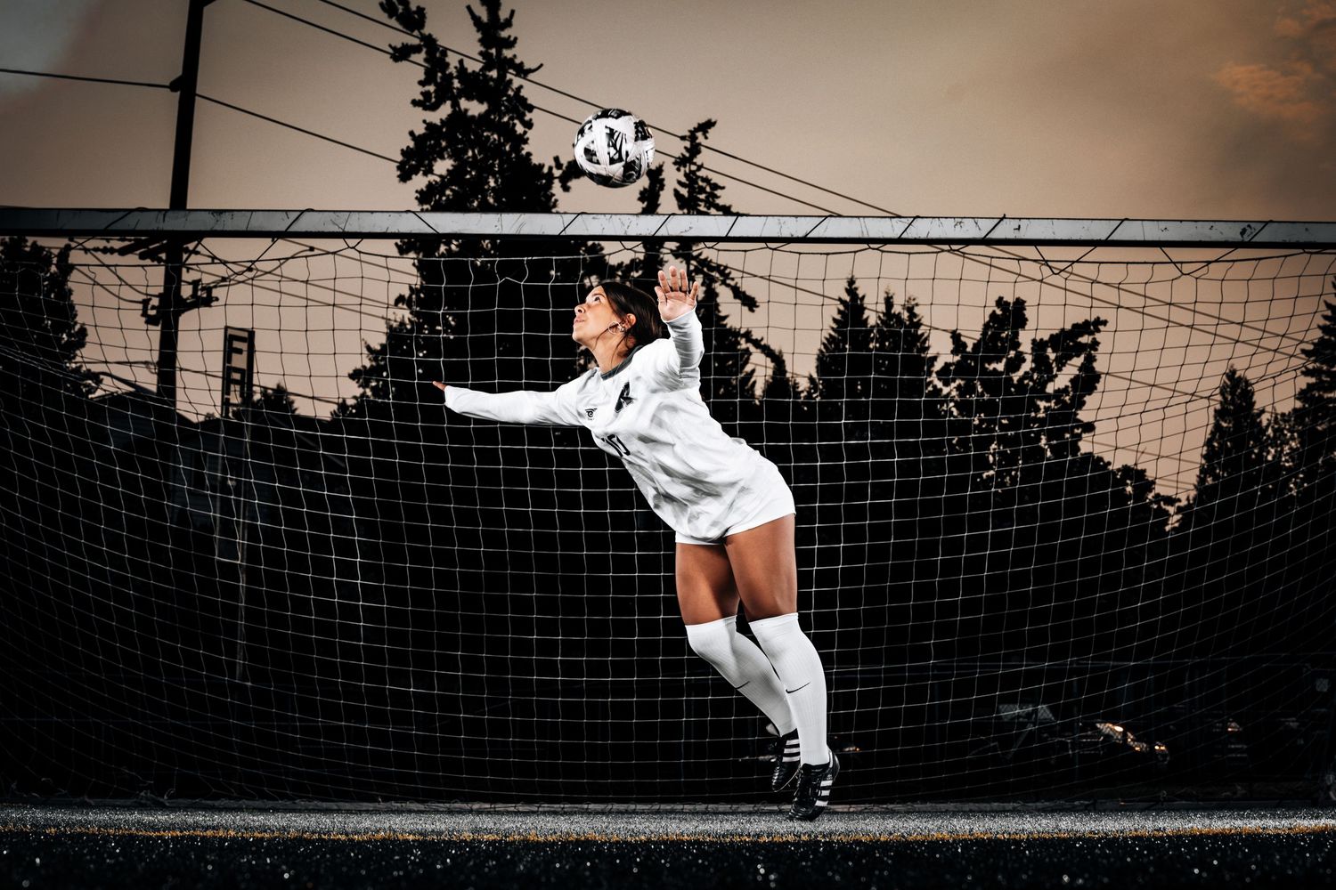 Soccer goalkeeper in white uniform makes a dramatic diving save attempt at sunset against dark silhouetted trees.