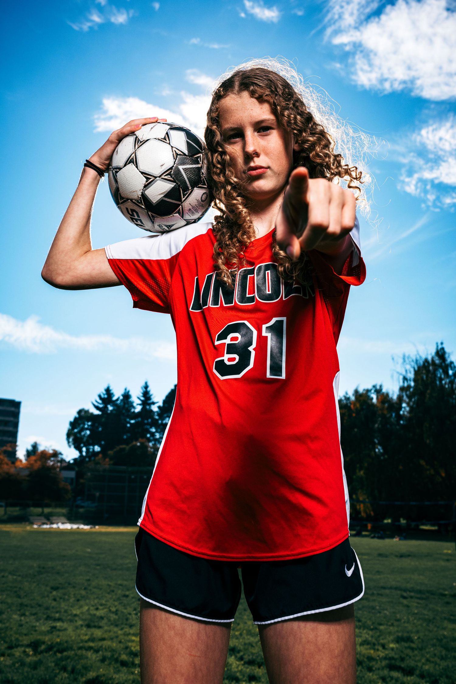 Soccer player in red Inco #31 jersey holds ball against blue sky with dramatic lighting and curly hair.