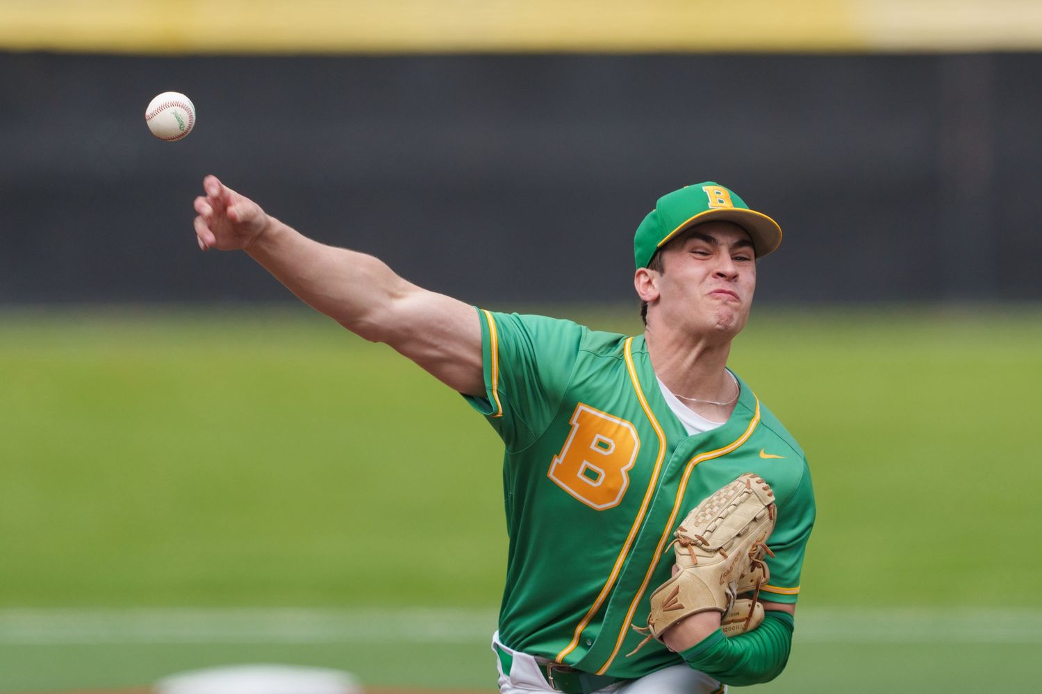 Baseball pitcher in green uniform throws pitch during game with ball visible mid-air against blurred background.