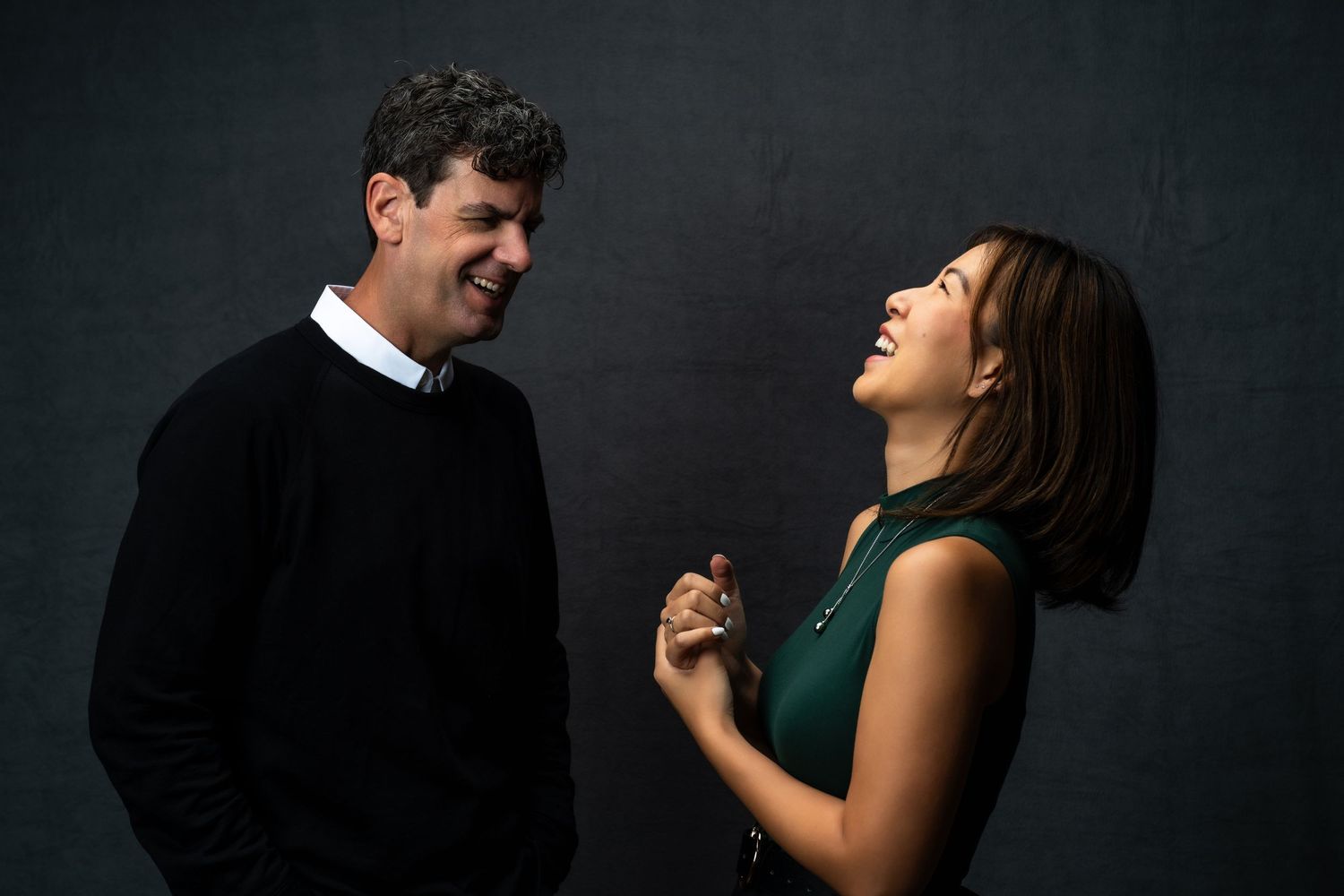Two people share a candid laughing moment together against a dark background during a portrait session.