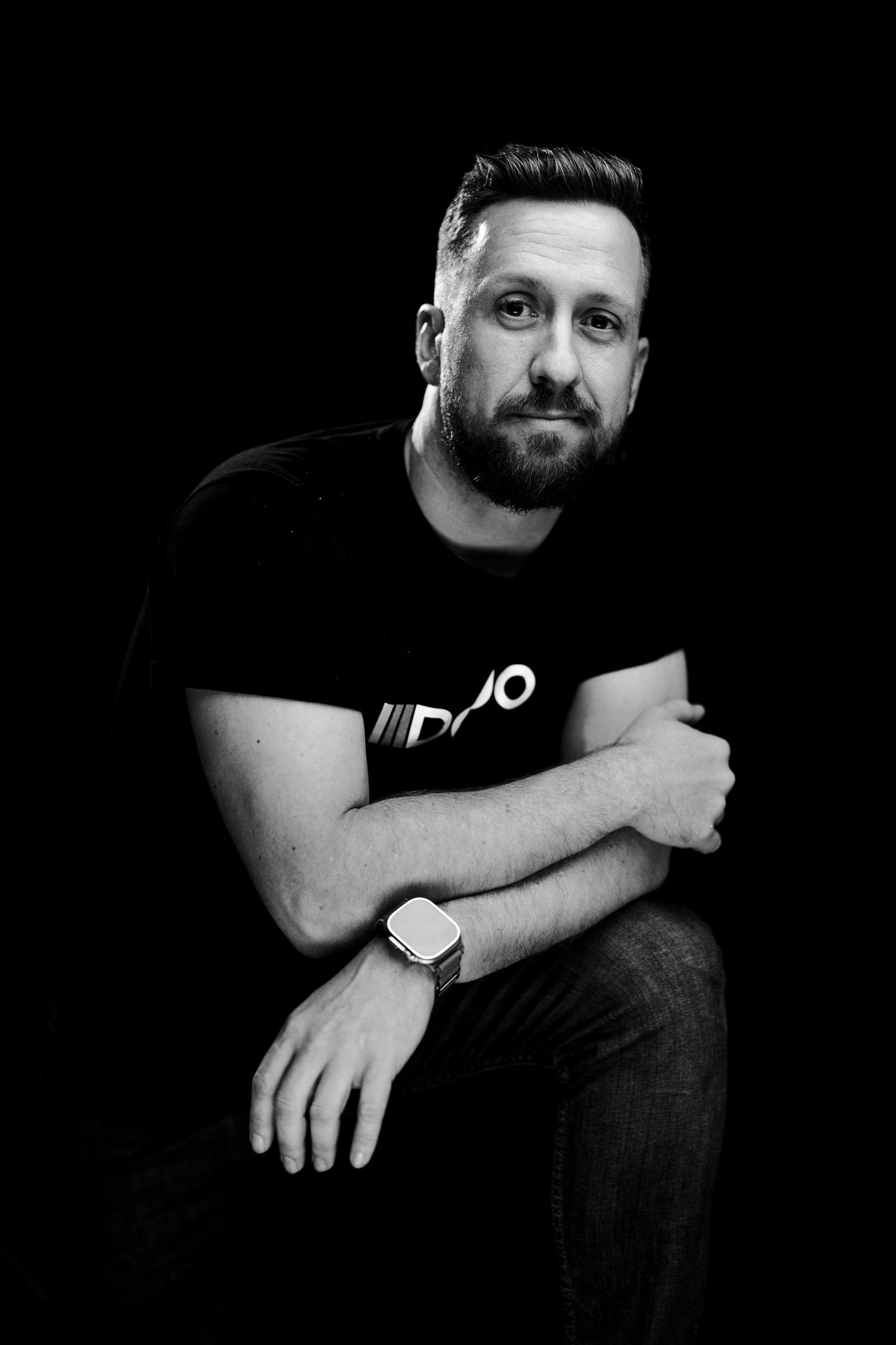Black and white portrait of person in dark t-shirt with crossed arms and wristwatch against black background.