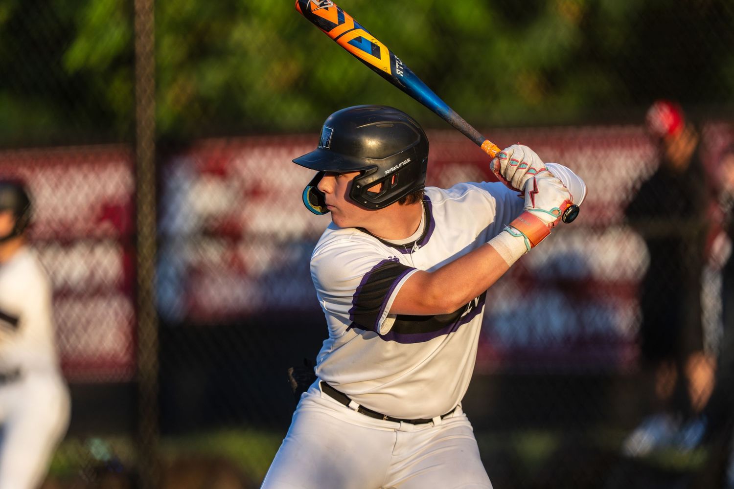 Baseball batter in white uniform and black helmet stands ready at home plate during evening game.