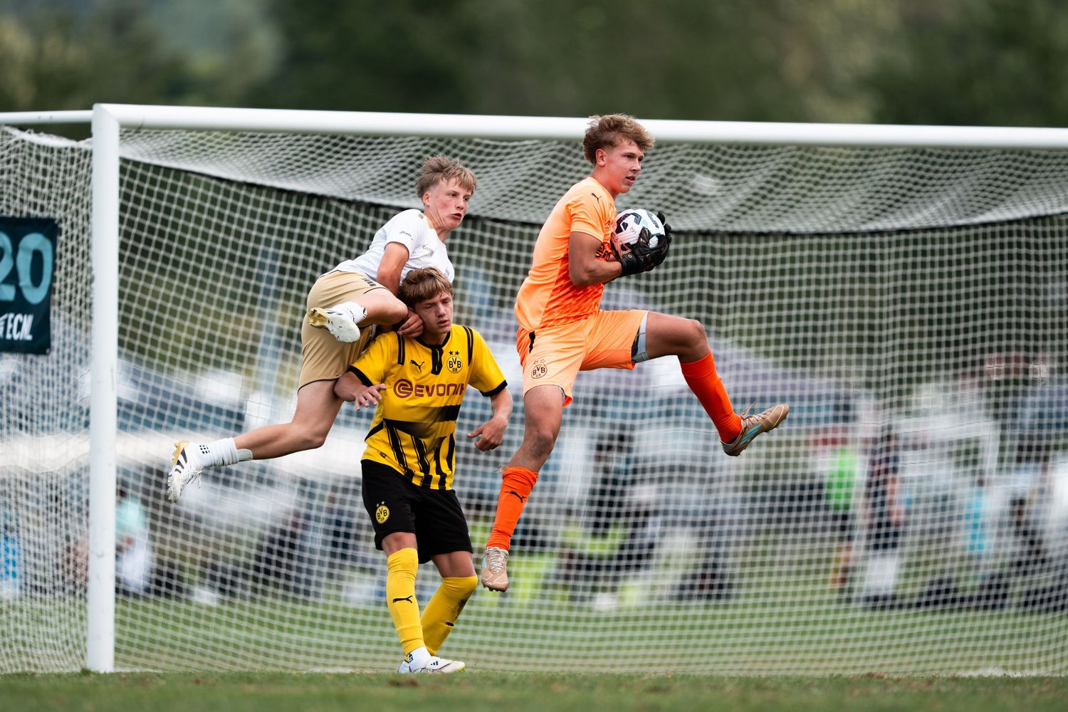Soccer goalkeeper in orange jersey makes a diving save against player in yellow and black striped uniform during match.