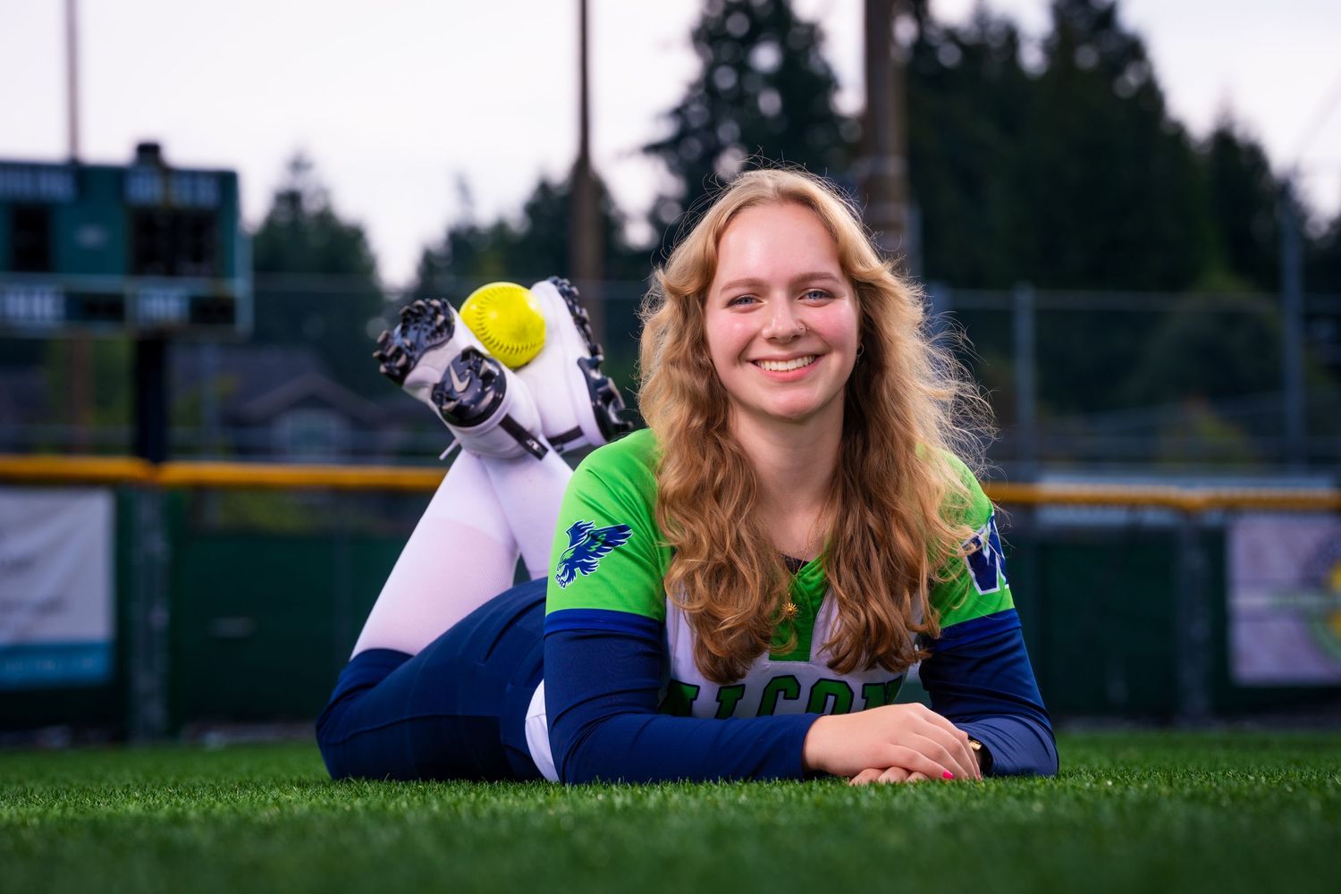 A person with wavy hair lies on grass while holding a yellow softball and smiling at the camera.