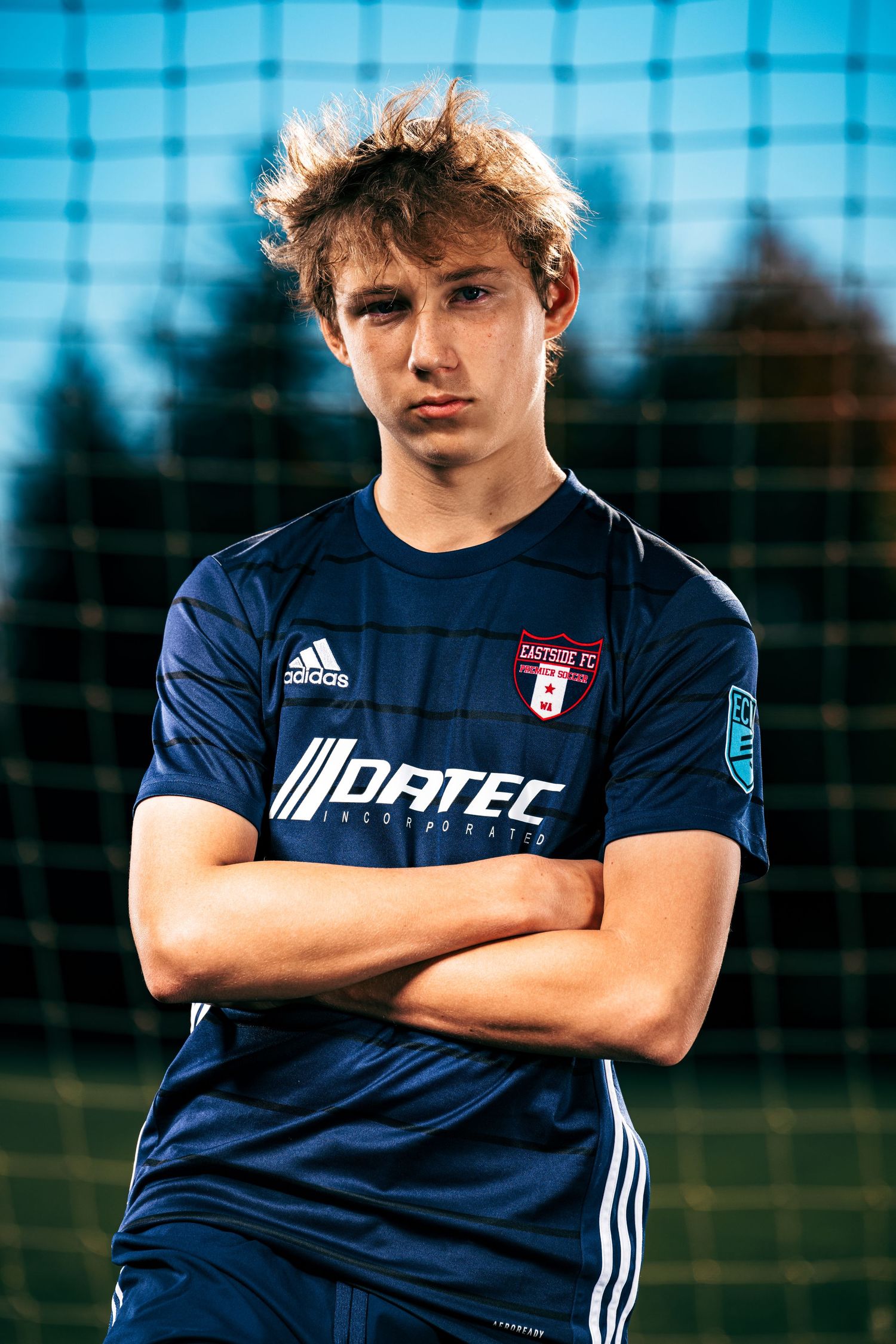 Soccer player wearing navy blue uniform with white stripes stands with arms crossed in outdoor setting.