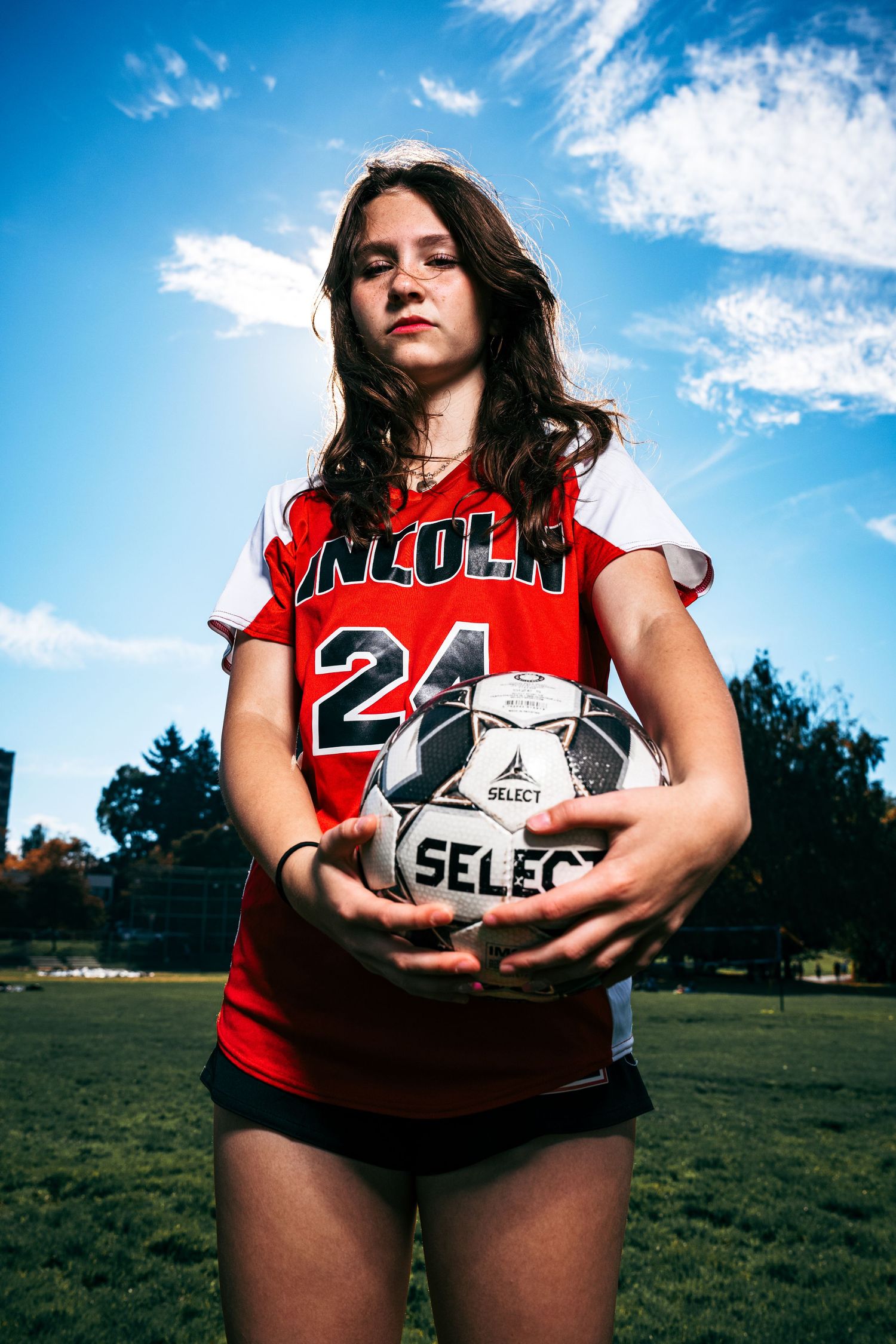 A soccer player in a red jersey holds a Select brand ball against a blue sky backdrop with trees in the background.
