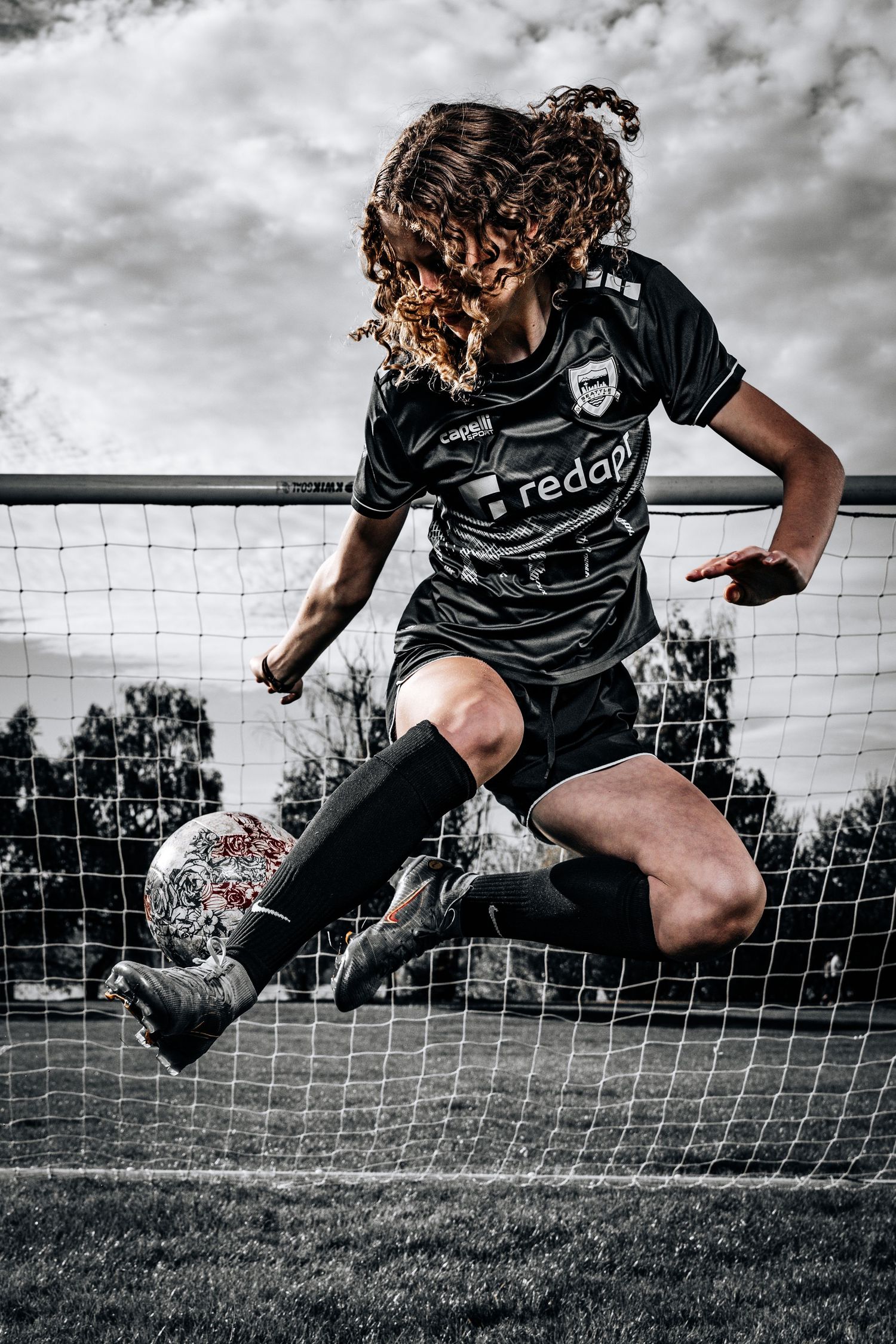 Dynamic soccer player in black Tedax uniform leaps to kick ball in front of goal net against cloudy sky.