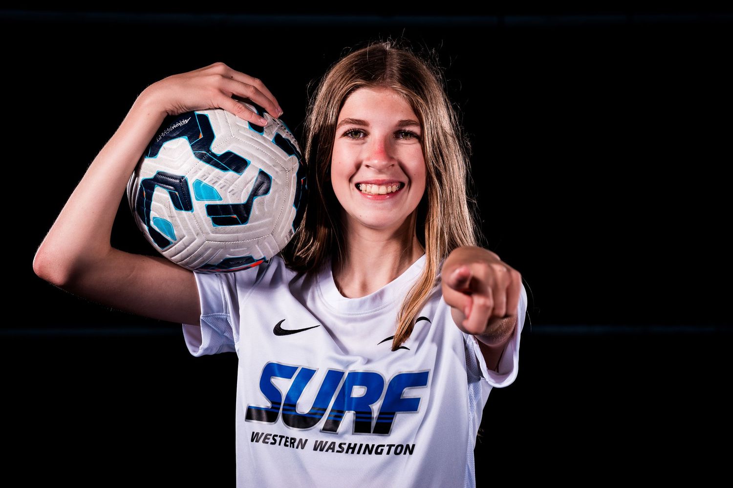 Soccer player in white jersey holds up ball while smiling at camera.