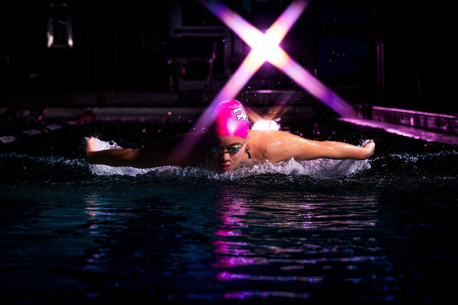 Synchronized swimmers perform a graceful split formation in dark water illuminated by dramatic purple stage lighting.