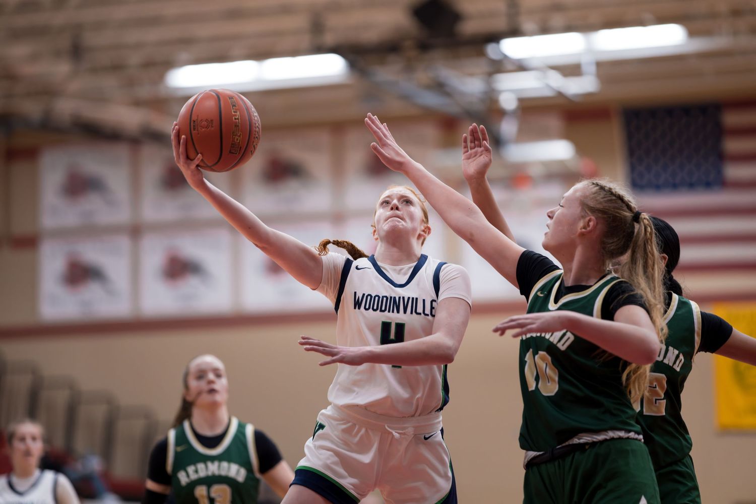 Two basketball players compete for the ball during a high school game with an American flag in the background.