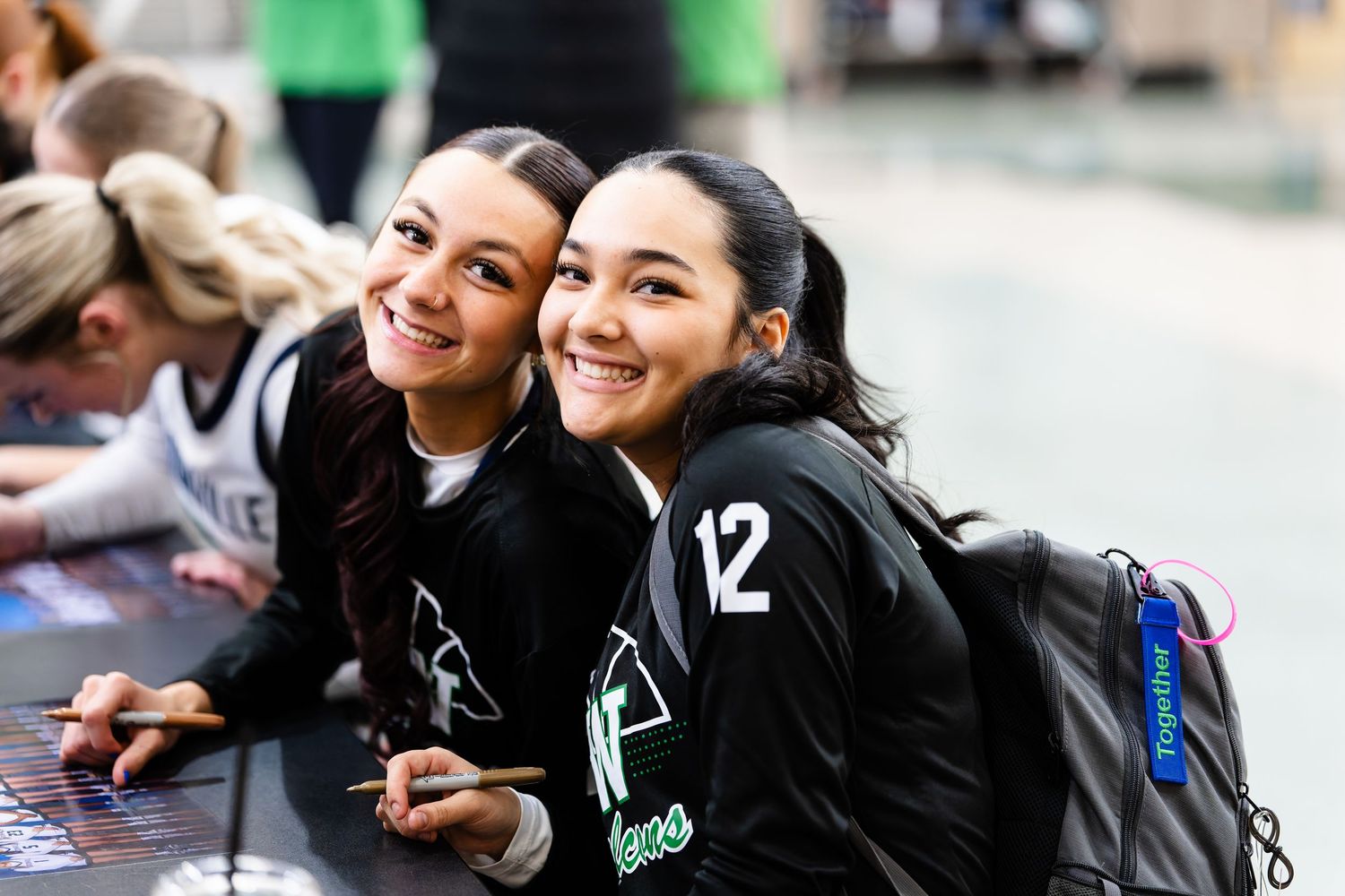 Two people in black jackets smile and laugh while sitting together at an outdoor event.