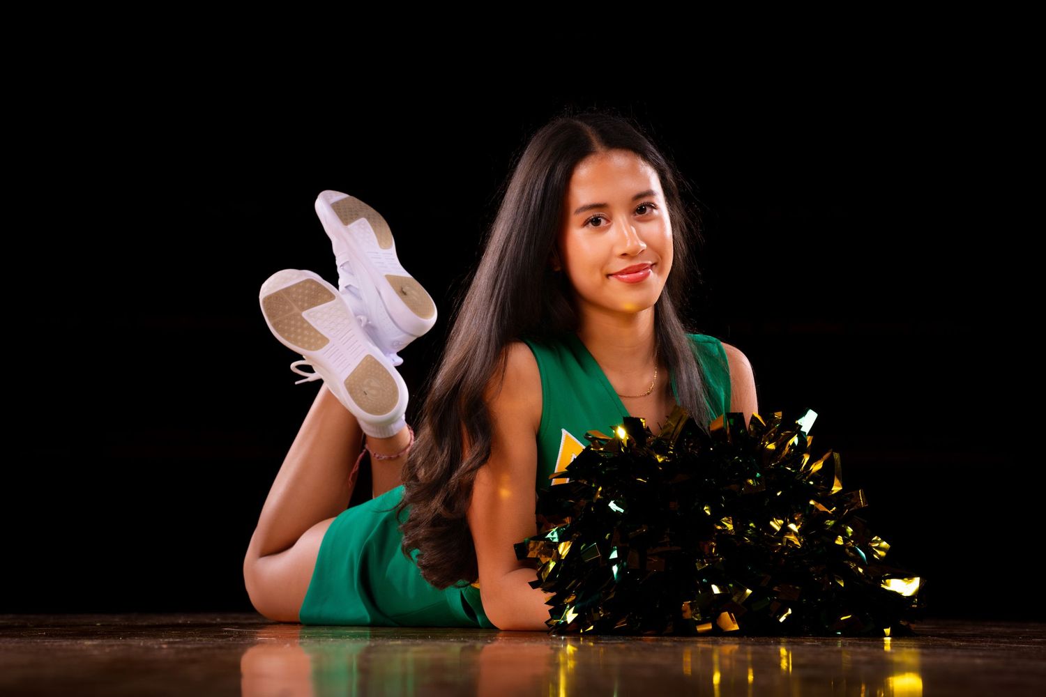 Cheerleader in green uniform with gold pom poms performing routine against dark background.