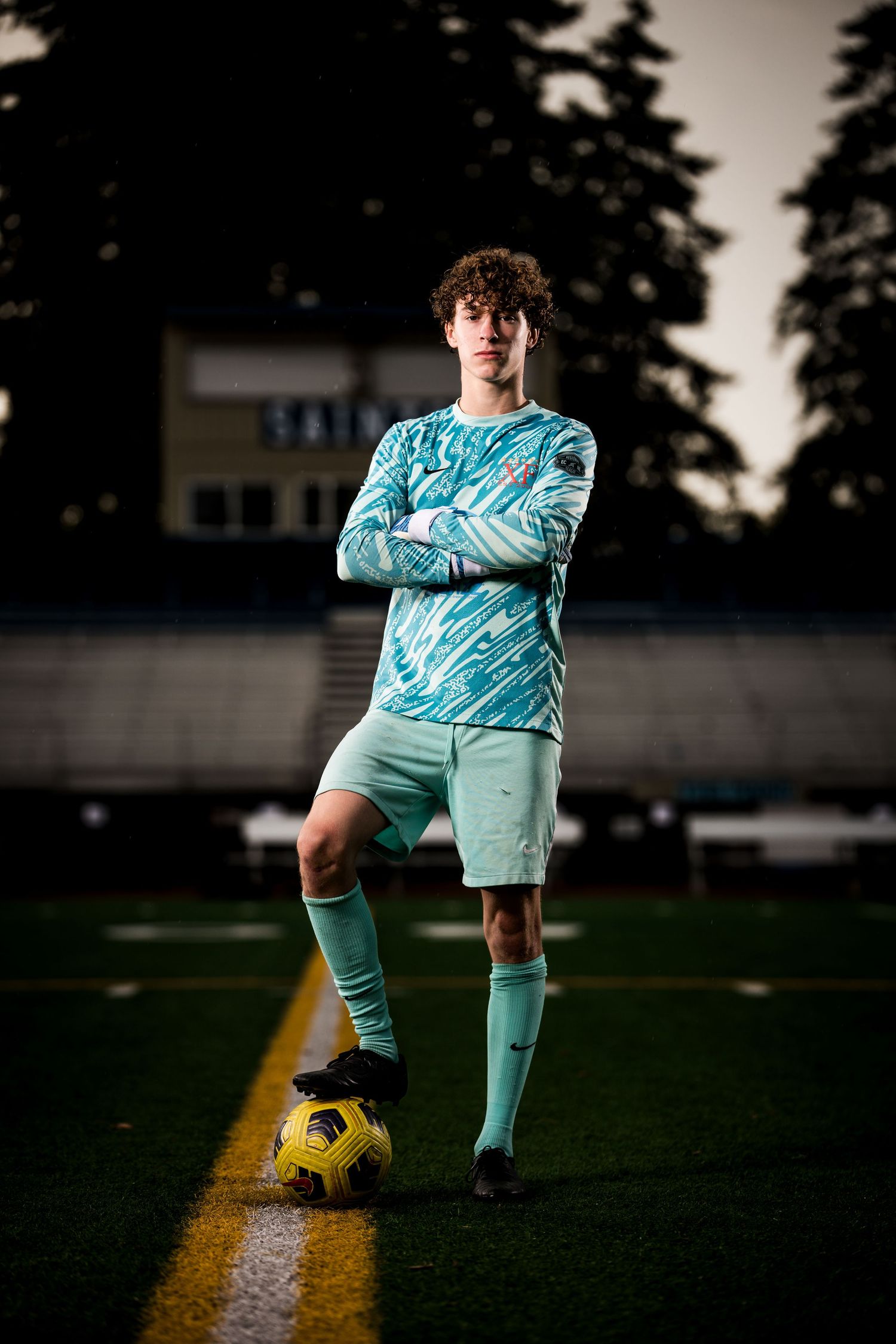 Soccer player in teal uniform poses with ball on field at dusk.