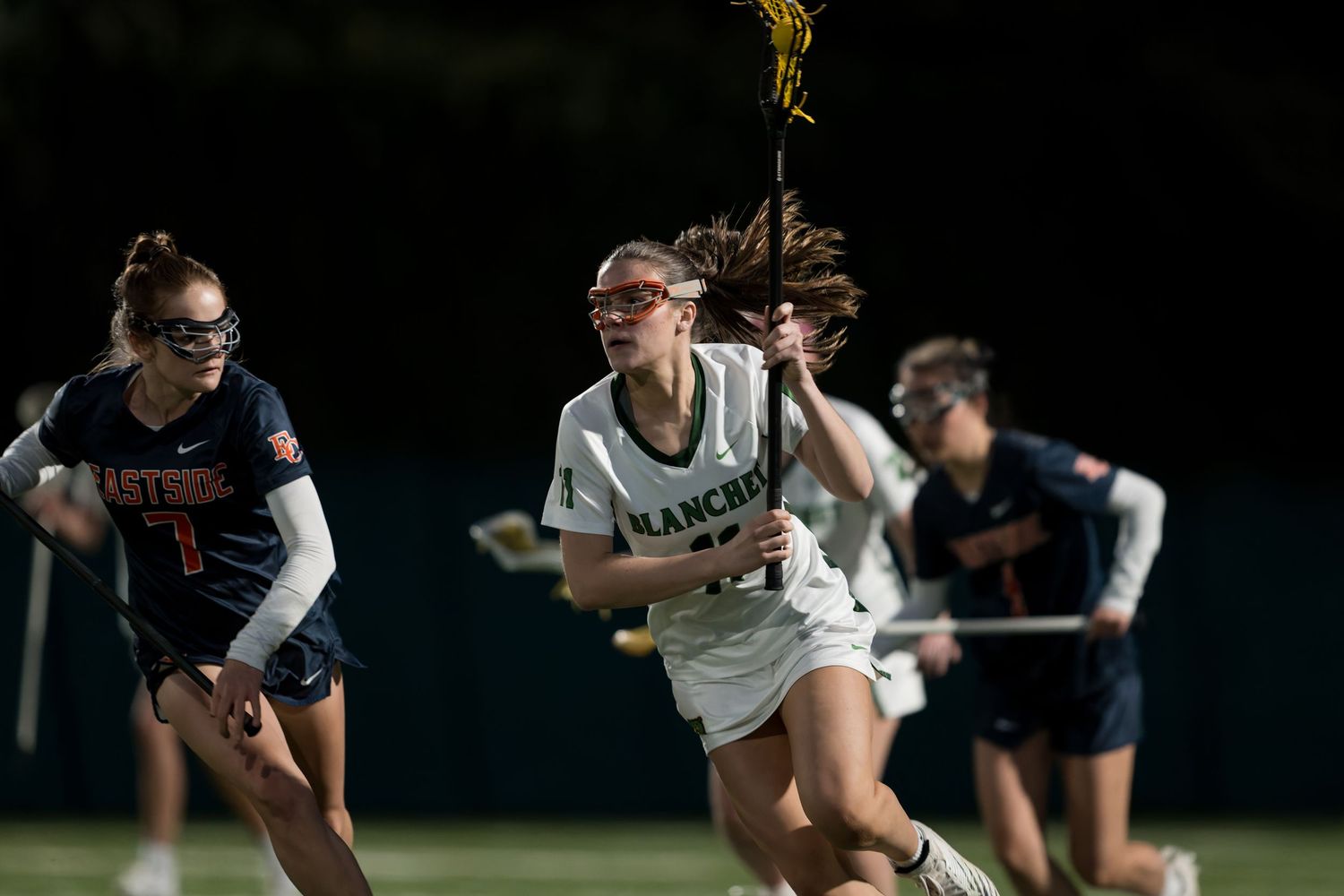 Lacrosse player in white jersey carries ball while running past defenders during nighttime game on field.
