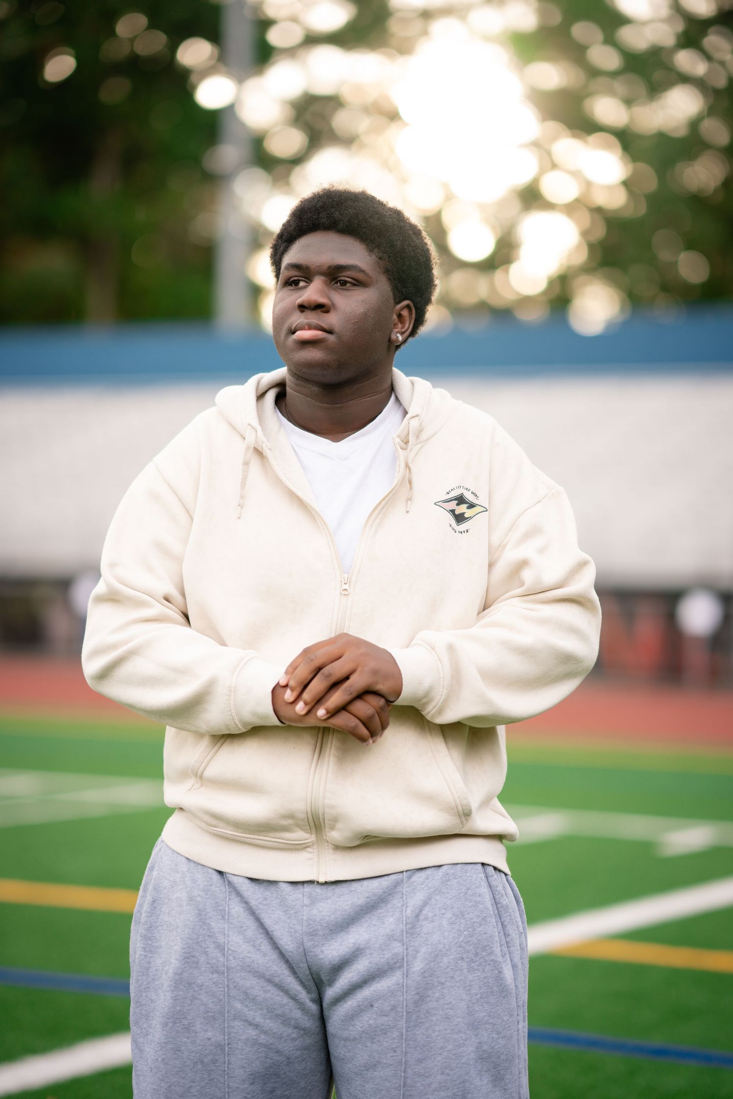A person wearing a cream colored sweatshirt and gray sweatpants sits outdoors on a sports field at dusk.
