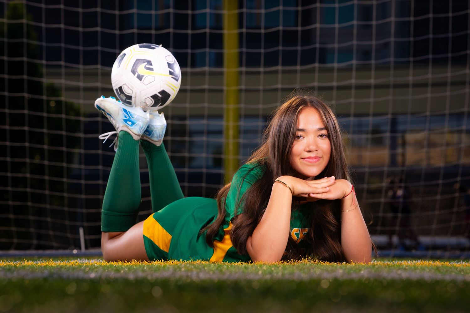 Soccer player in green uniform lying on grass field with soccer ball during sports action shot.