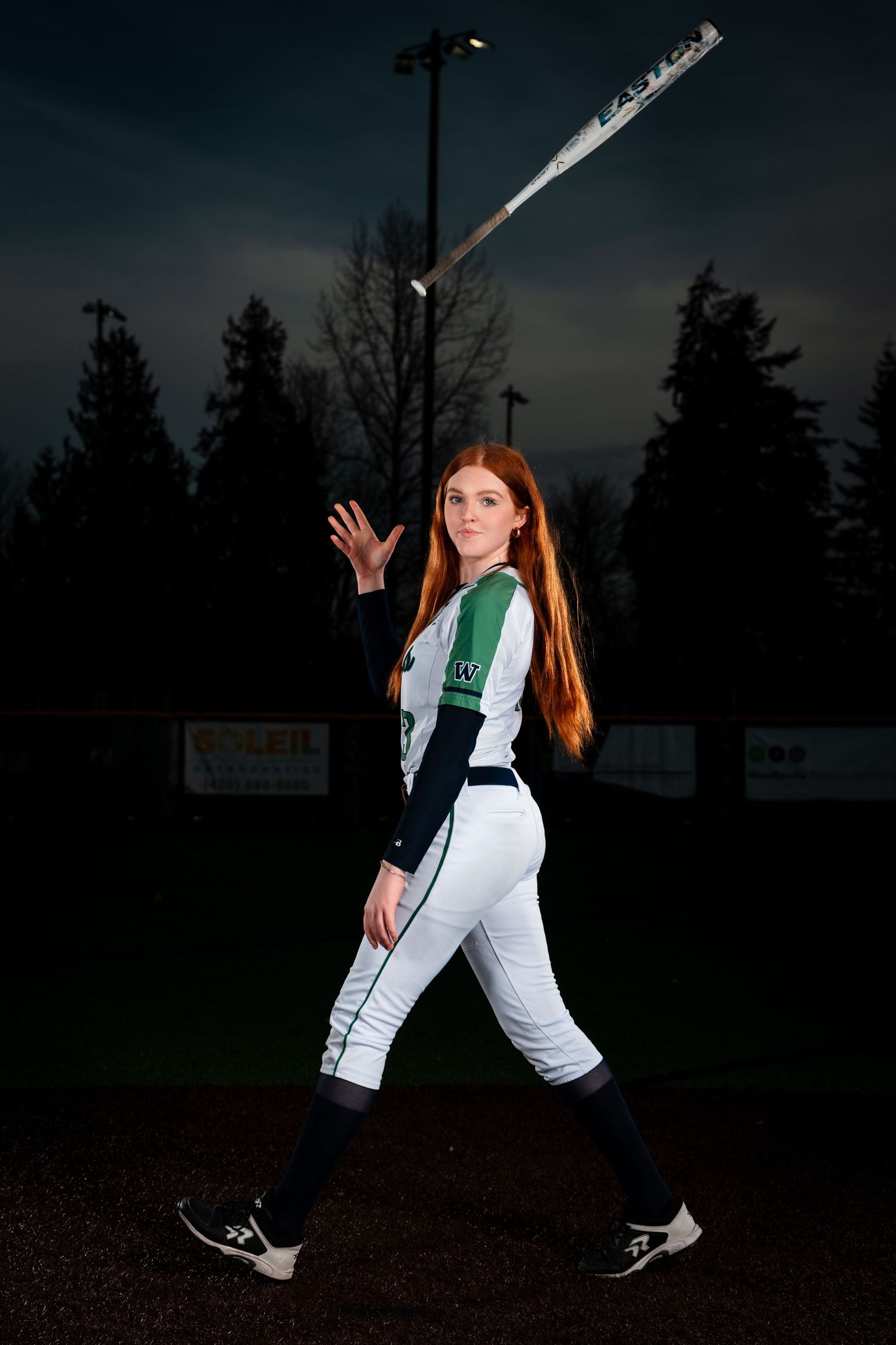 Softball player in white and green uniform poses at dusk with baseball bat under stadium lights.