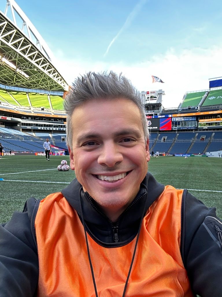 Person wearing orange safety vest smiling while standing on football field at stadium.