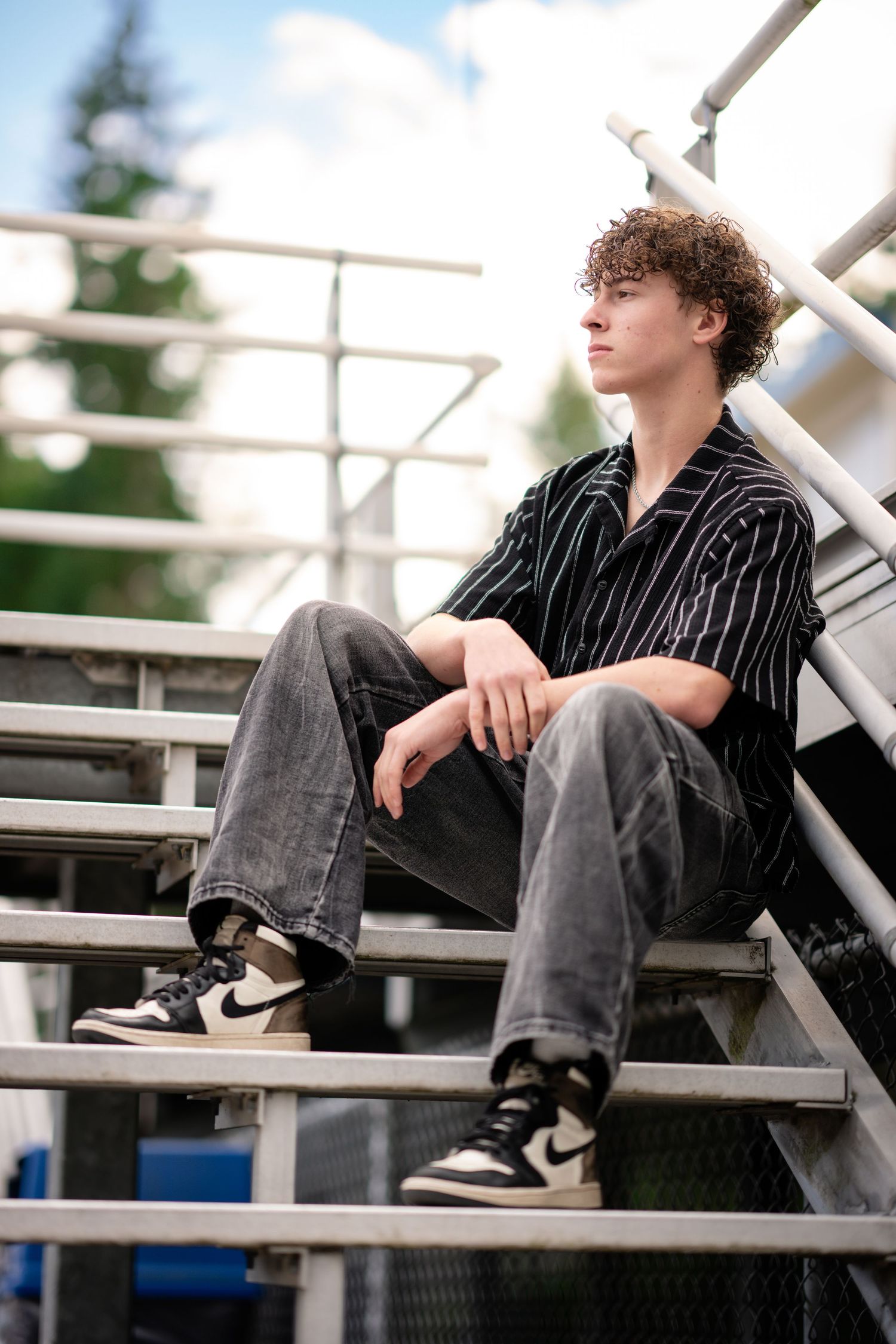 A person in a striped shirt and jeans sits casually on outdoor metal bleachers in a contemplative pose.