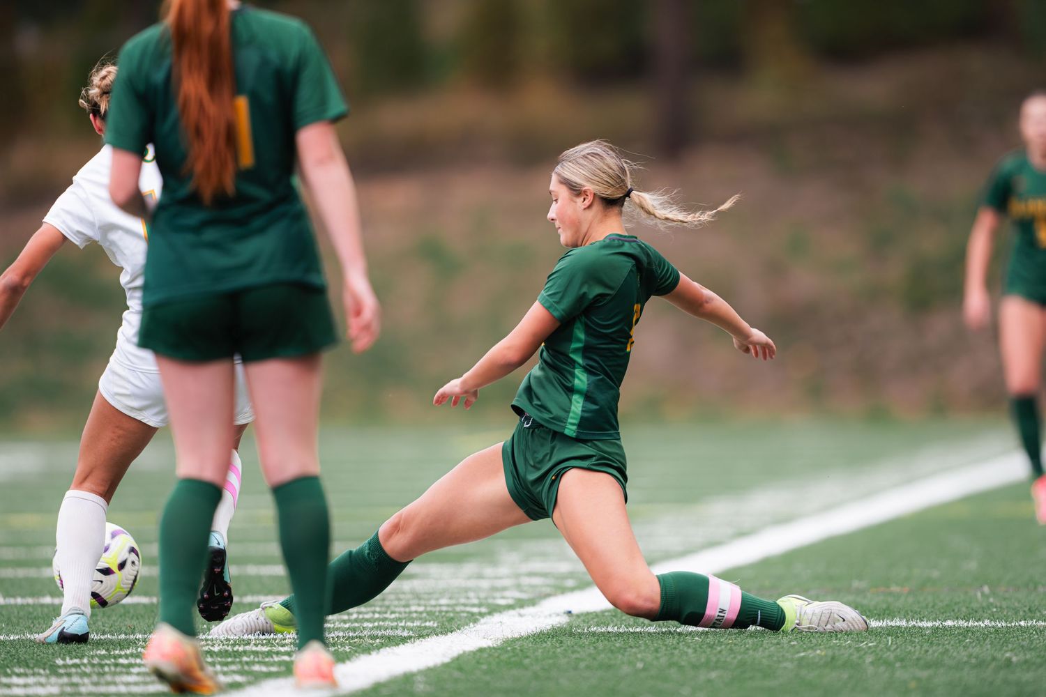 Soccer players in green and white uniforms slide tackle during competitive match on turf field.