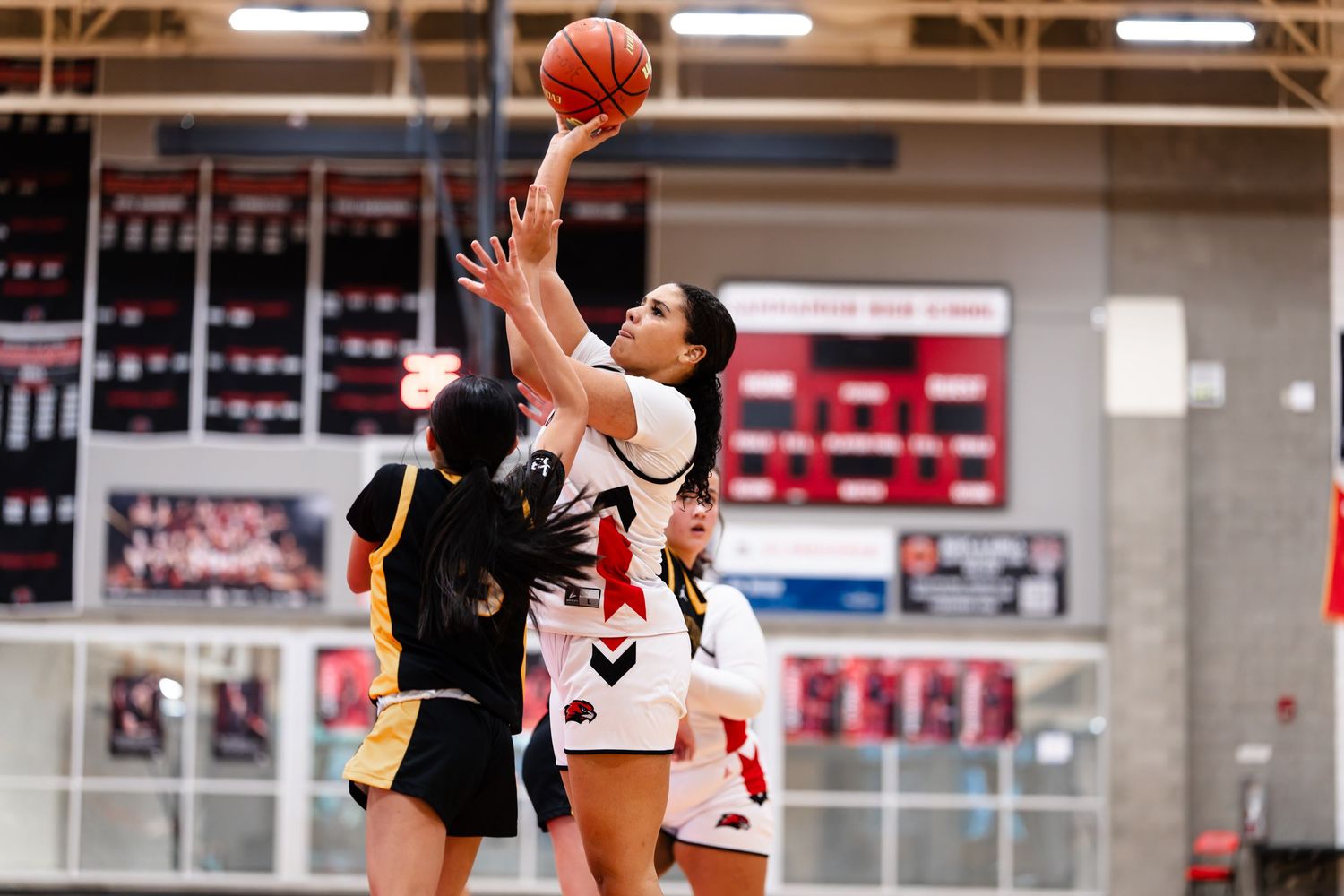 Basketball players leap for a rebound during an intense game in a gymnasium with red seats visible in the background.