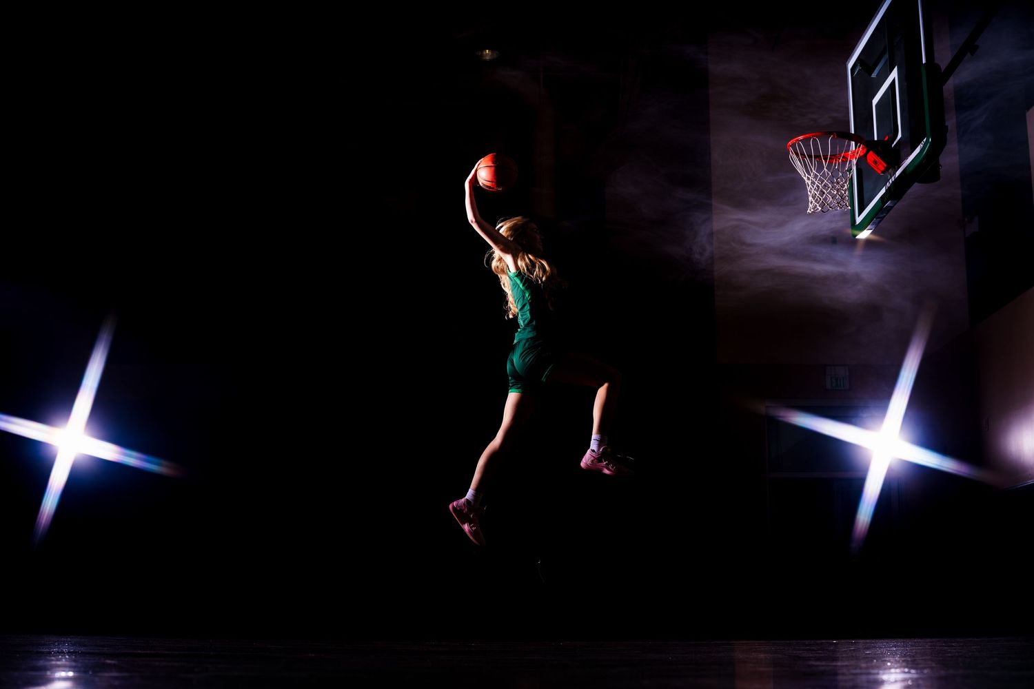 Basketball player silhouetted against spotlights on dark court with dramatic lighting.