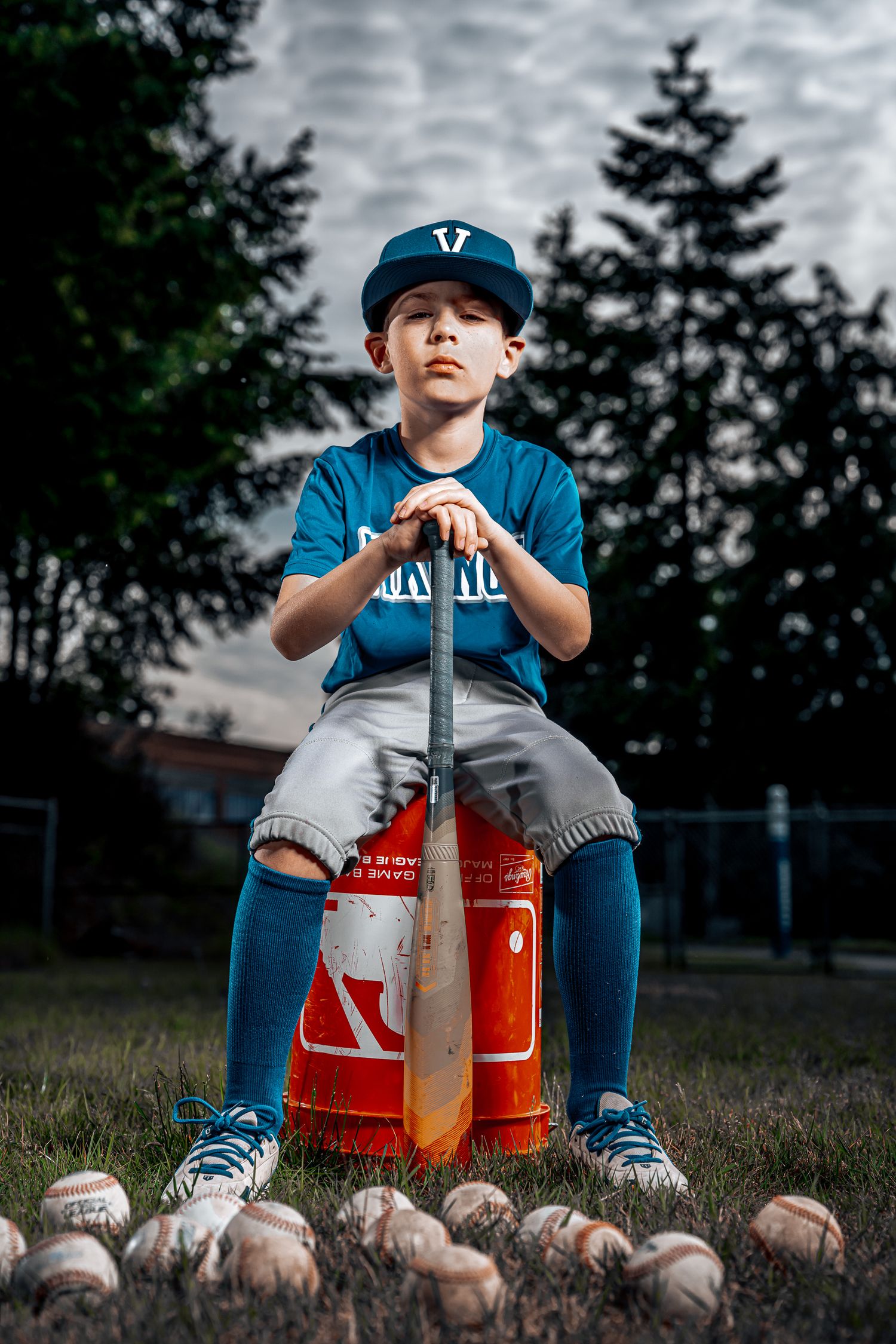 Youth baseball player in blue uniform sits on red bucket surrounded by baseballs at dusk.