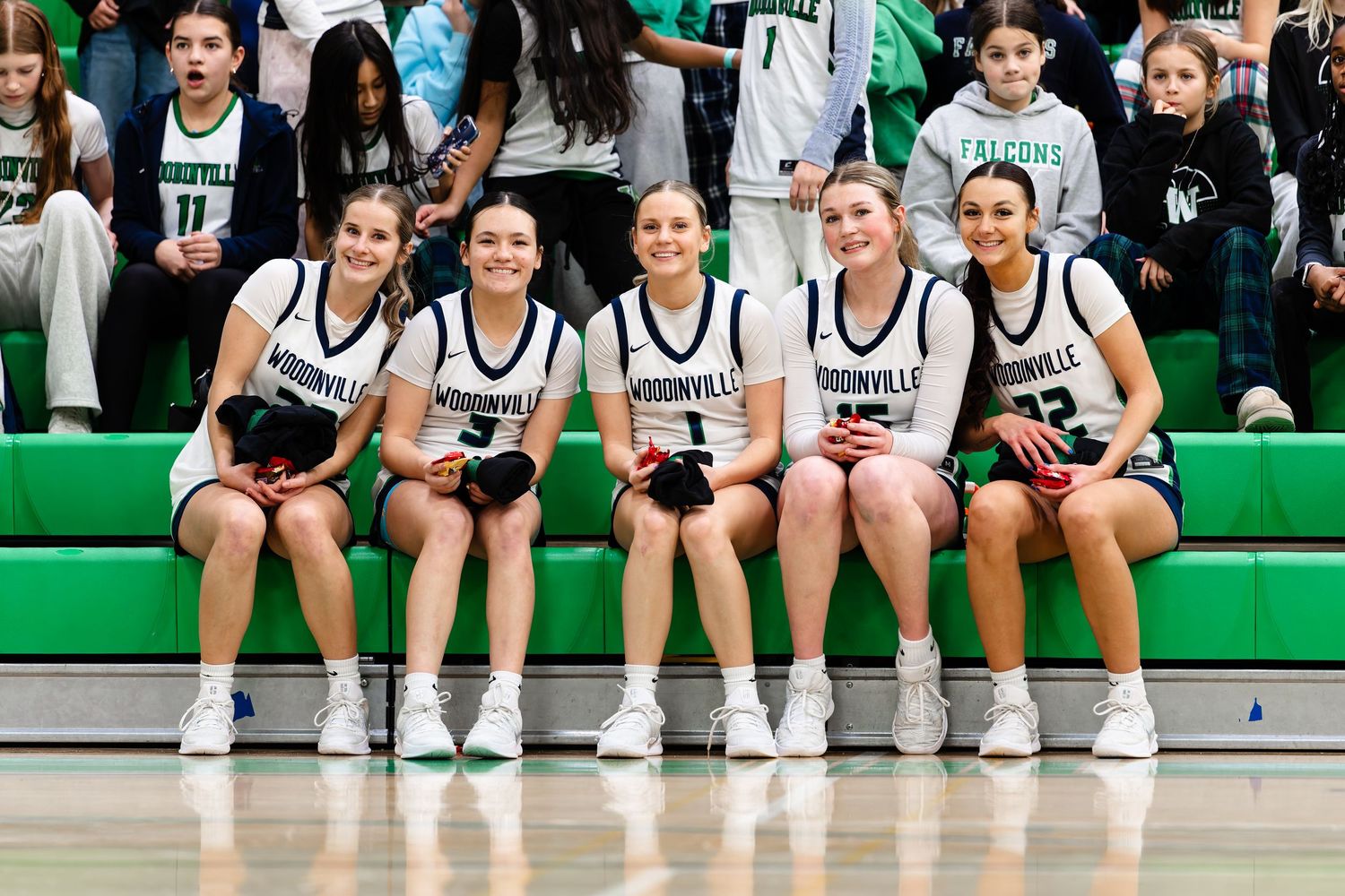 Basketball players in white and black uniforms sit together on bench during game at green-colored venue.