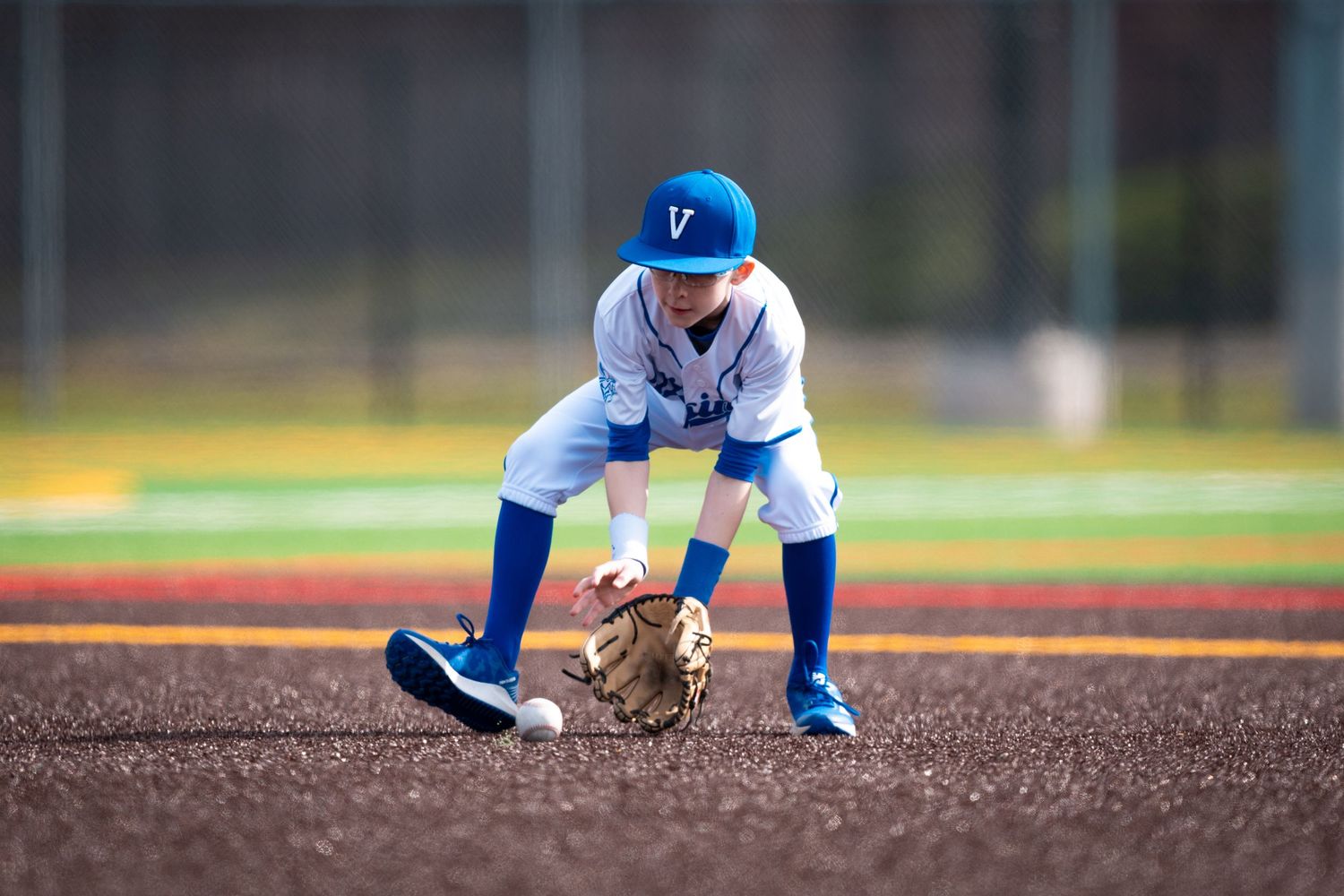 Youth baseball player in blue and white uniform crouches in fielding position on infield dirt during practice.