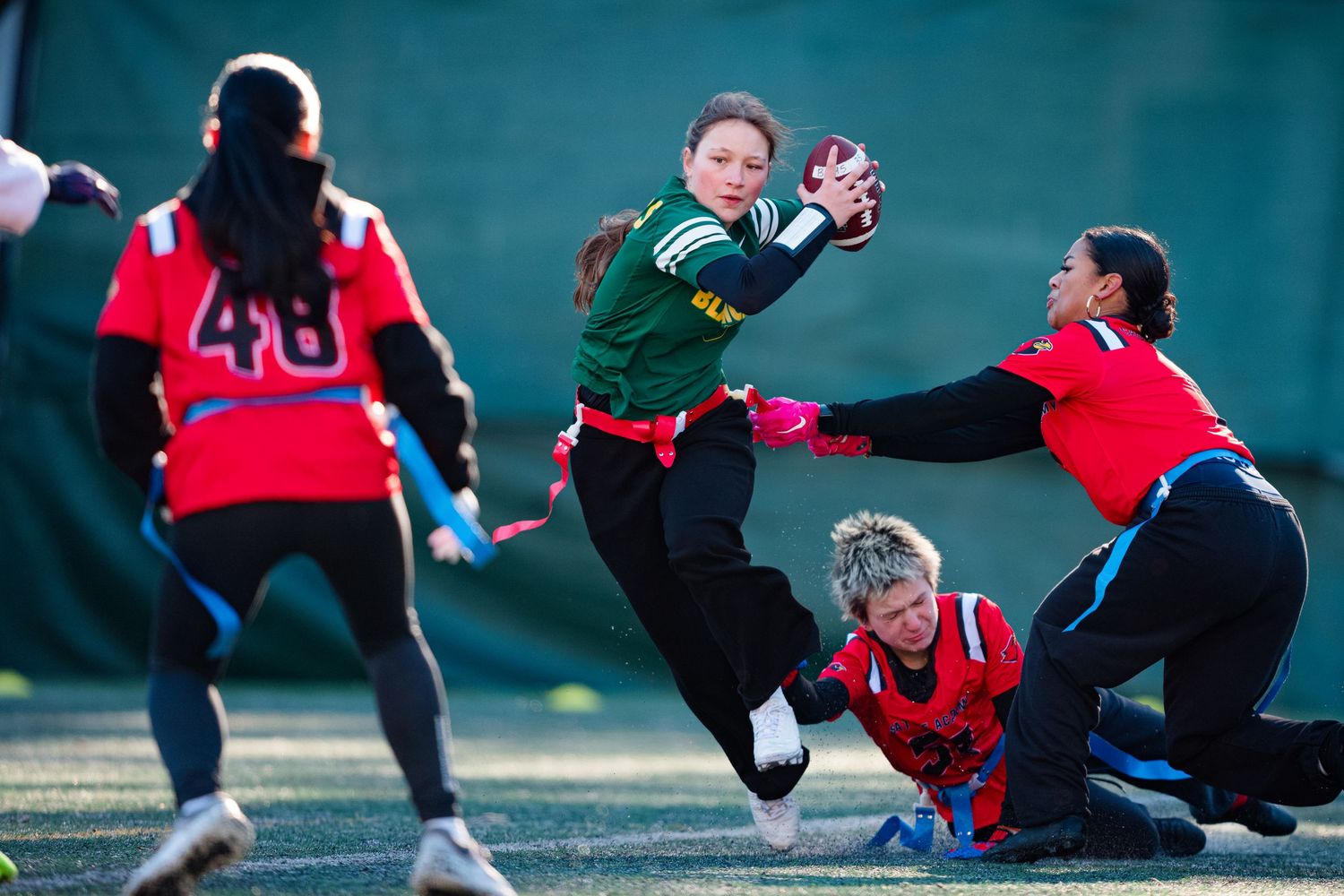 Soccer players slide tackle during an intense match on a grass field in red and black uniforms.