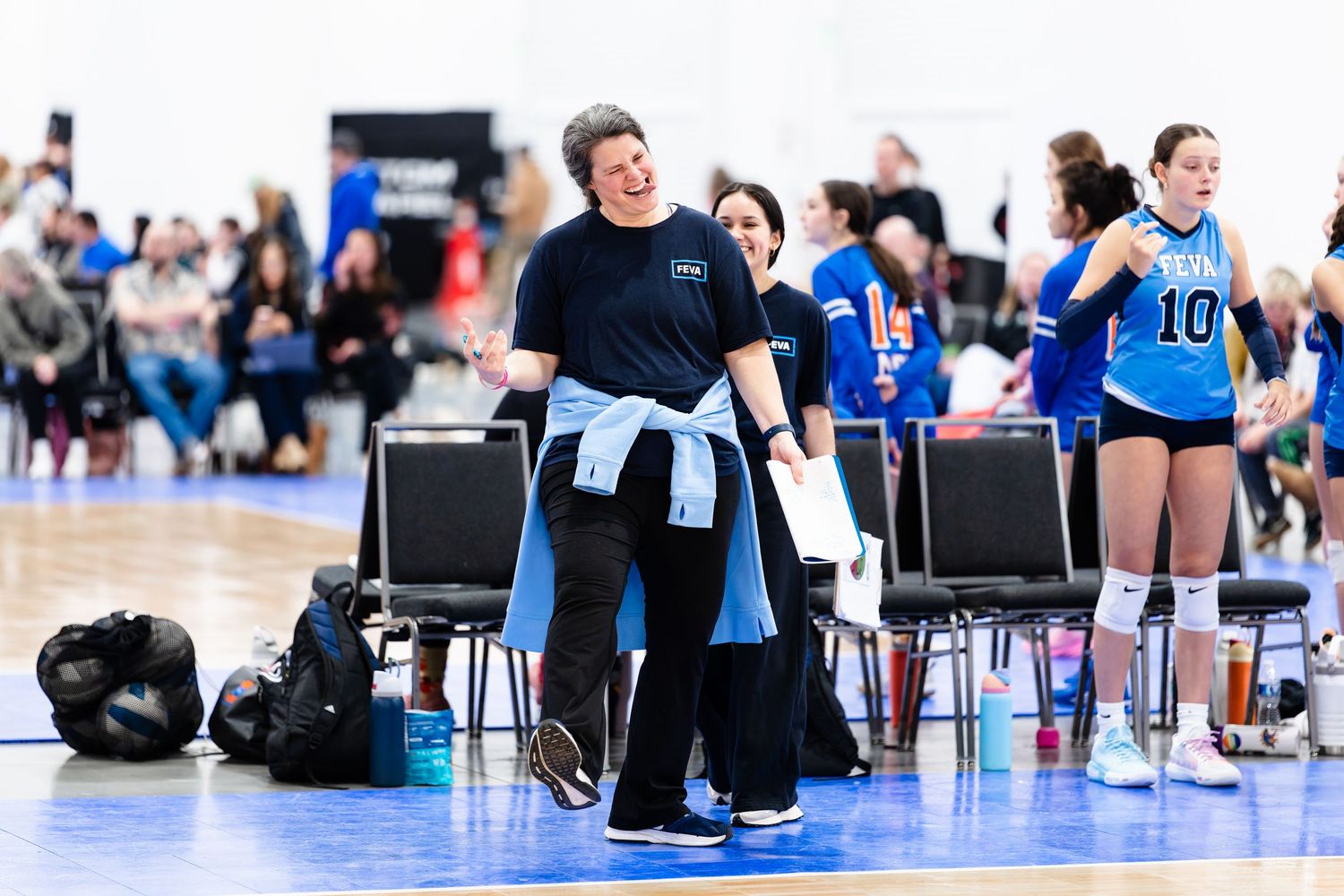 Athletes compete in an indoor sitting volleyball match on a blue court with spectators watching from the sidelines.