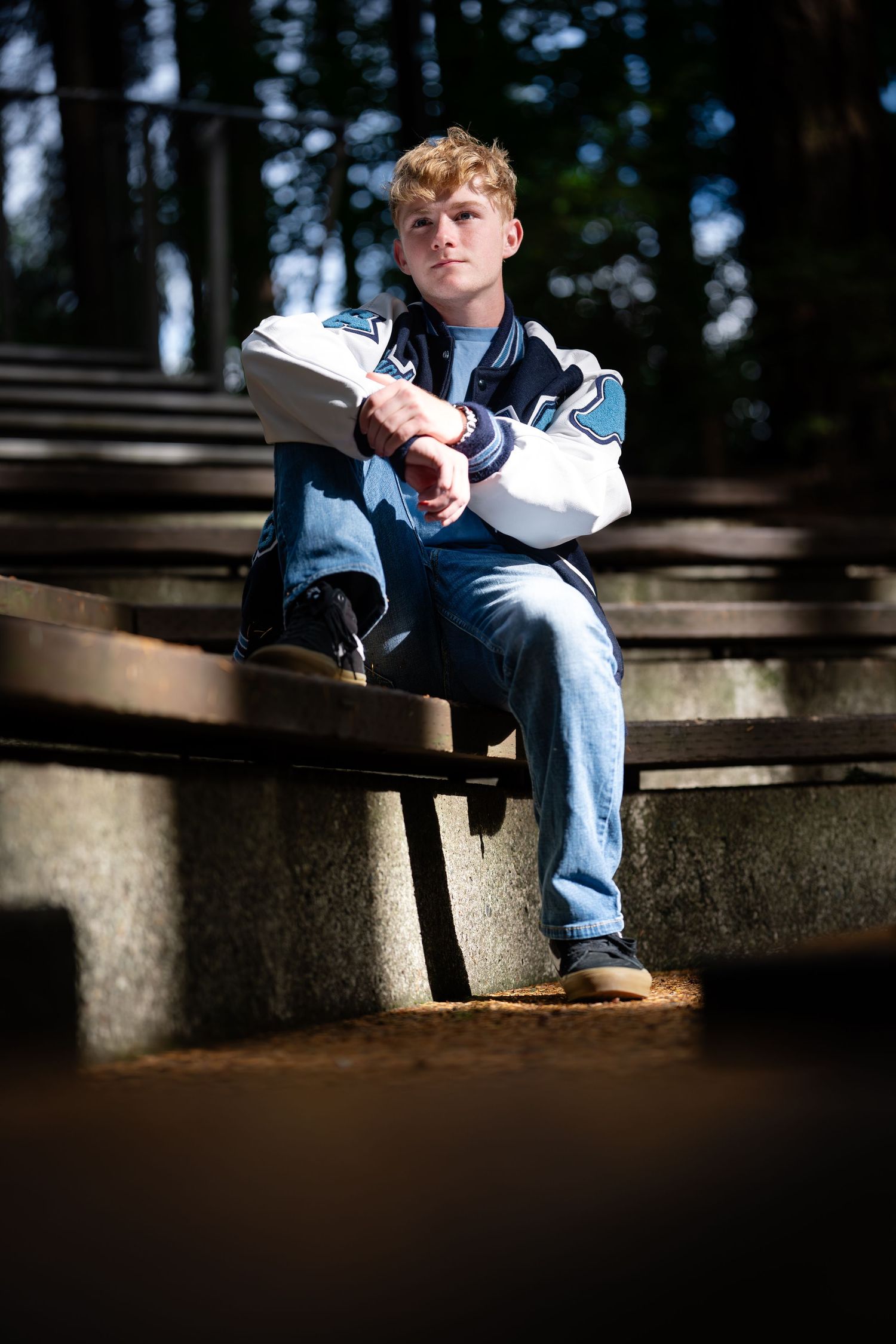 Someone sits on concrete steps in a wooded area wearing blue jeans and a navy jacket on a sunny day.