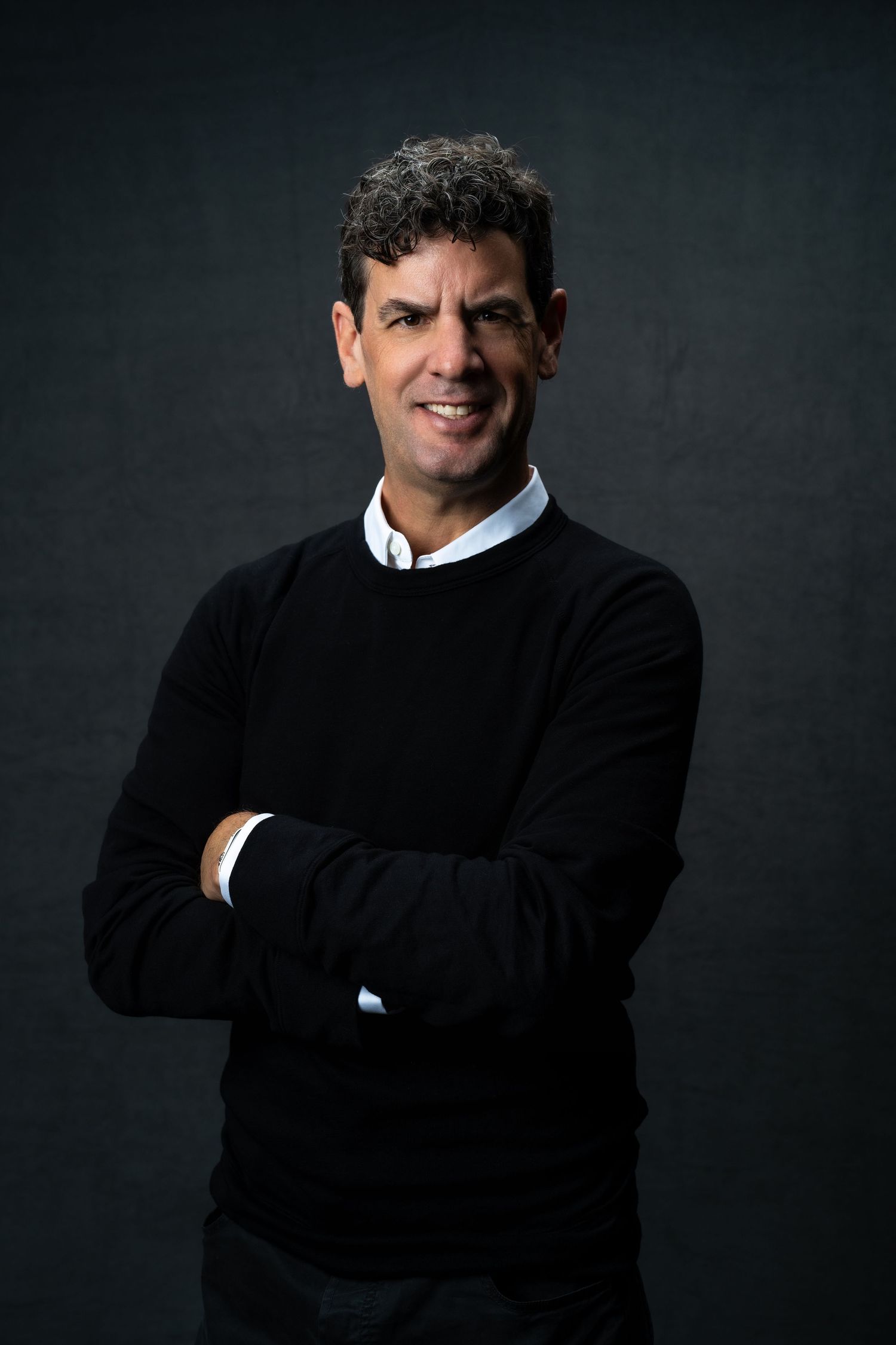 Professional studio portrait against dark background showing person in black sweater with crossed arms and confident pose.