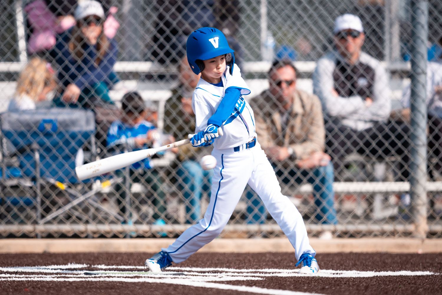 Baseball player in blue and white uniform swings bat during game with spectators watching through chain link fence.