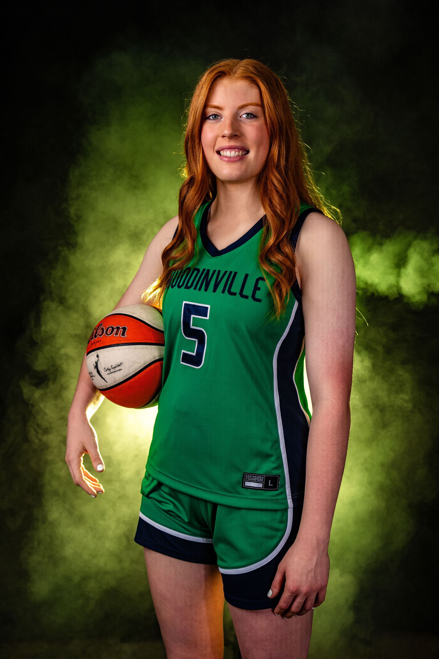 Basketball player in green Connell jersey poses with ball against dramatic dark background with green smoke effects.