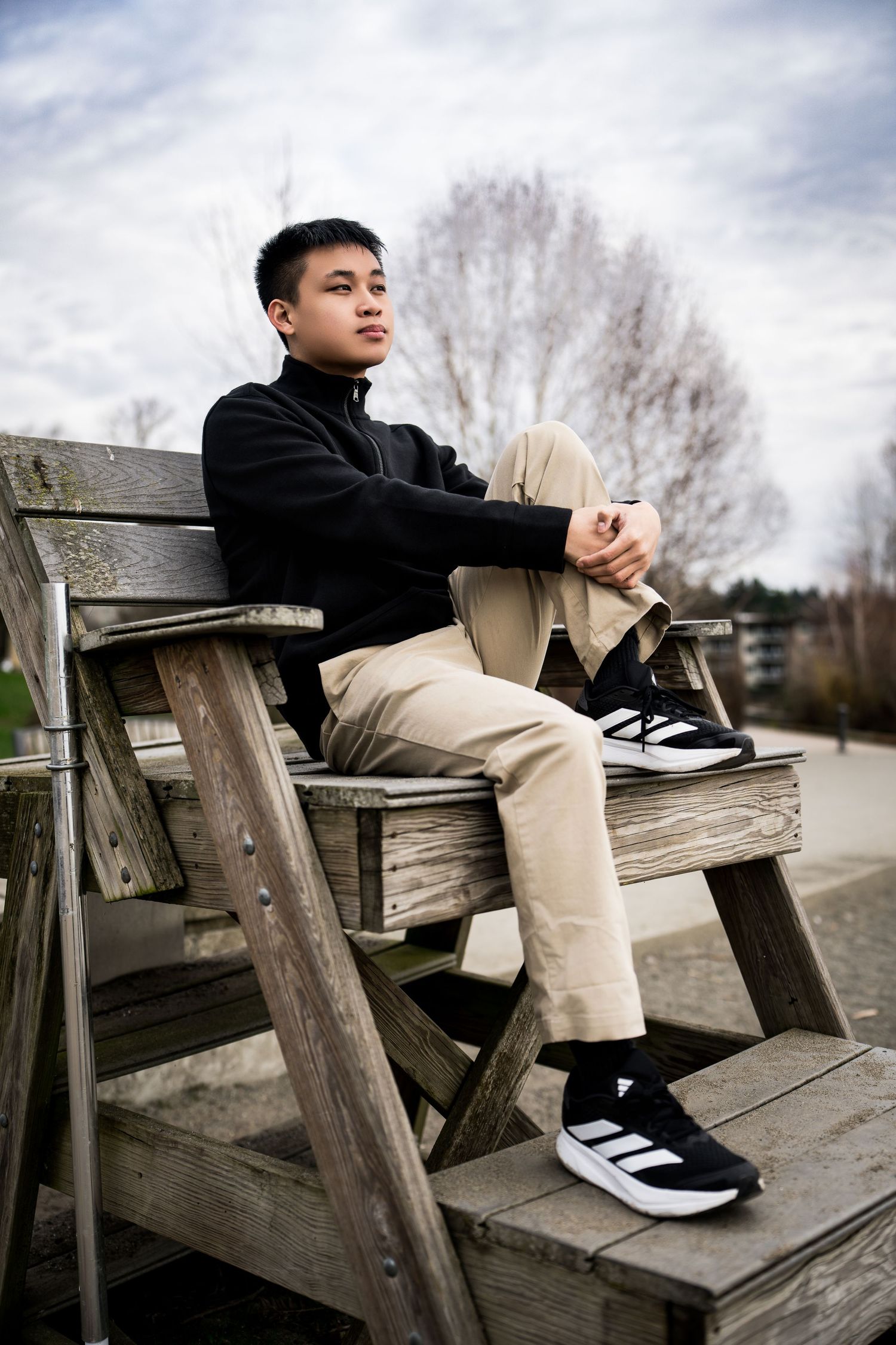Portrait of someone in black sweater and khaki pants sitting on wooden deck stairs on cloudy day.