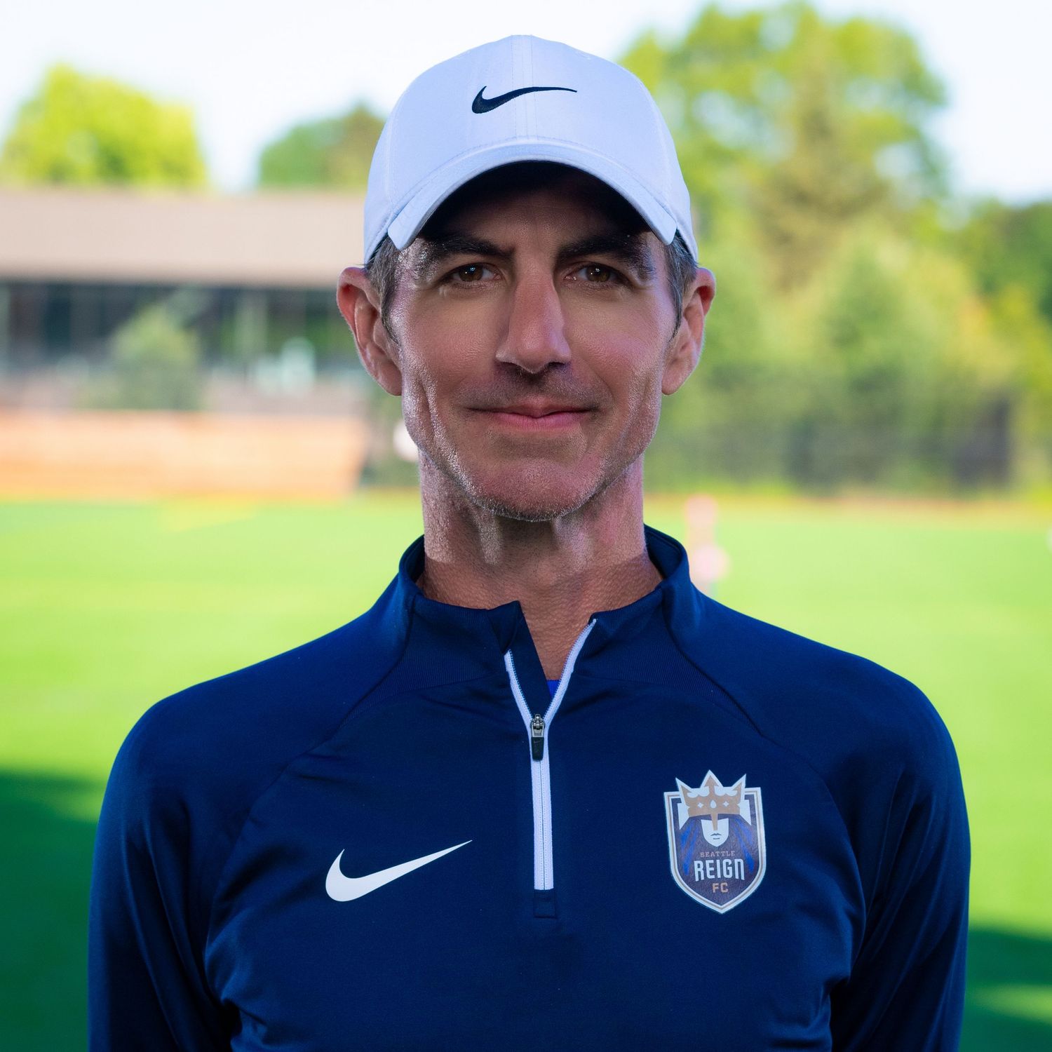 A coach wearing a Nike baseball cap and navy blue quarter-zip jacket stands on a sunny sports field.