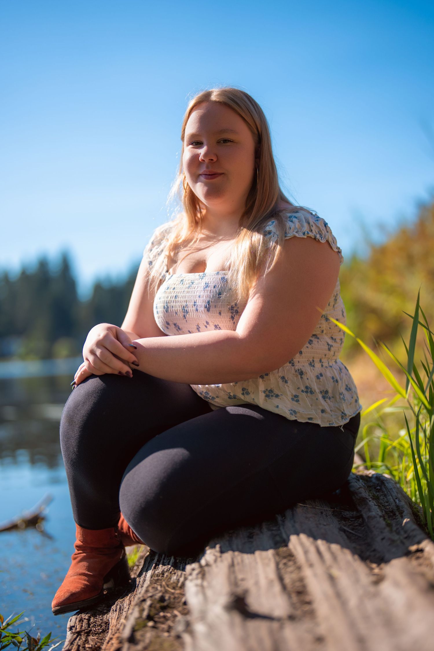 A person in a floral dress sits by a lake on a sunny day with water and trees in the background.