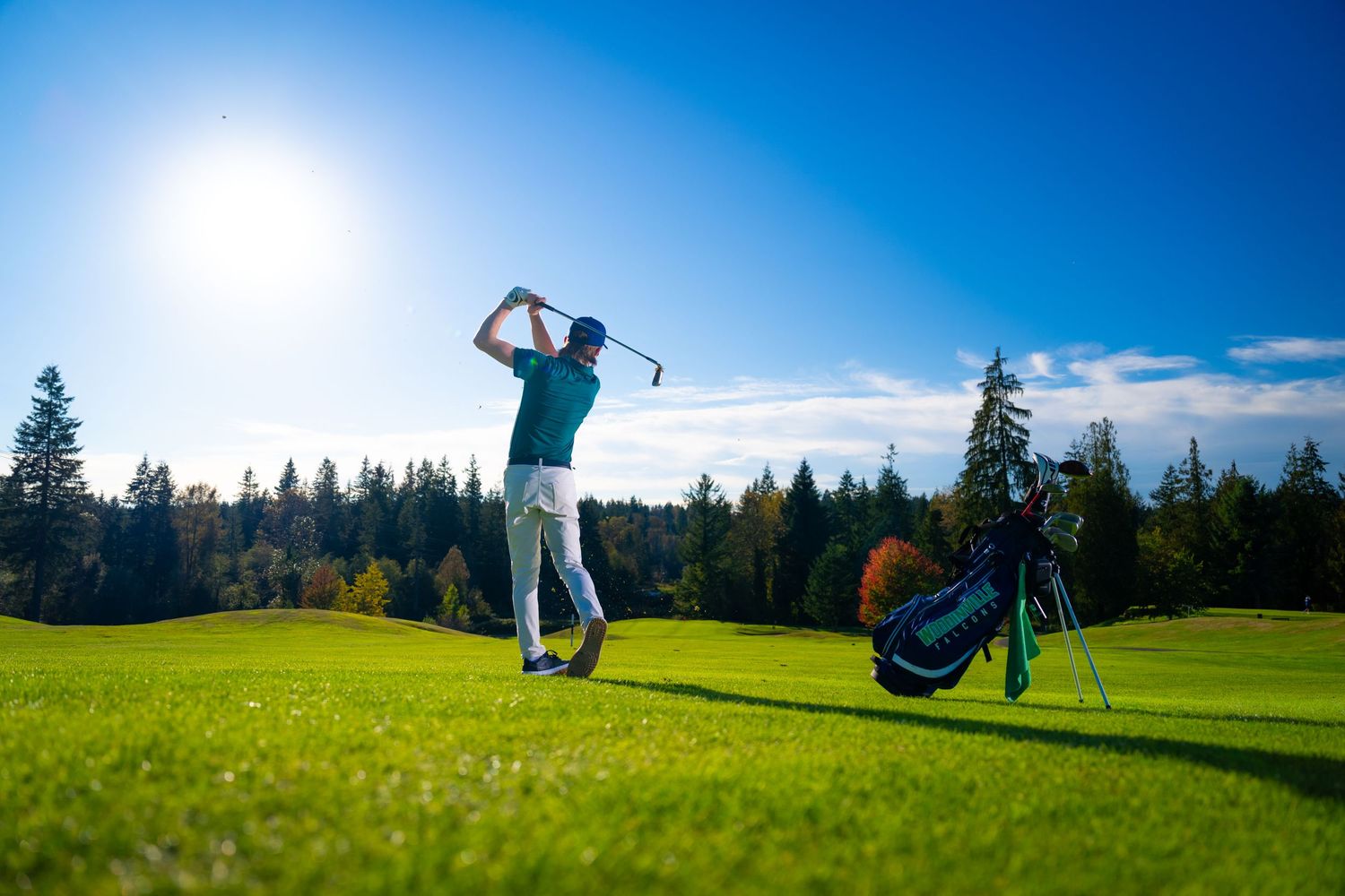 Golfer taking a swing on a lush green course with golf bag and evergreen forest backdrop on a sunny day.
