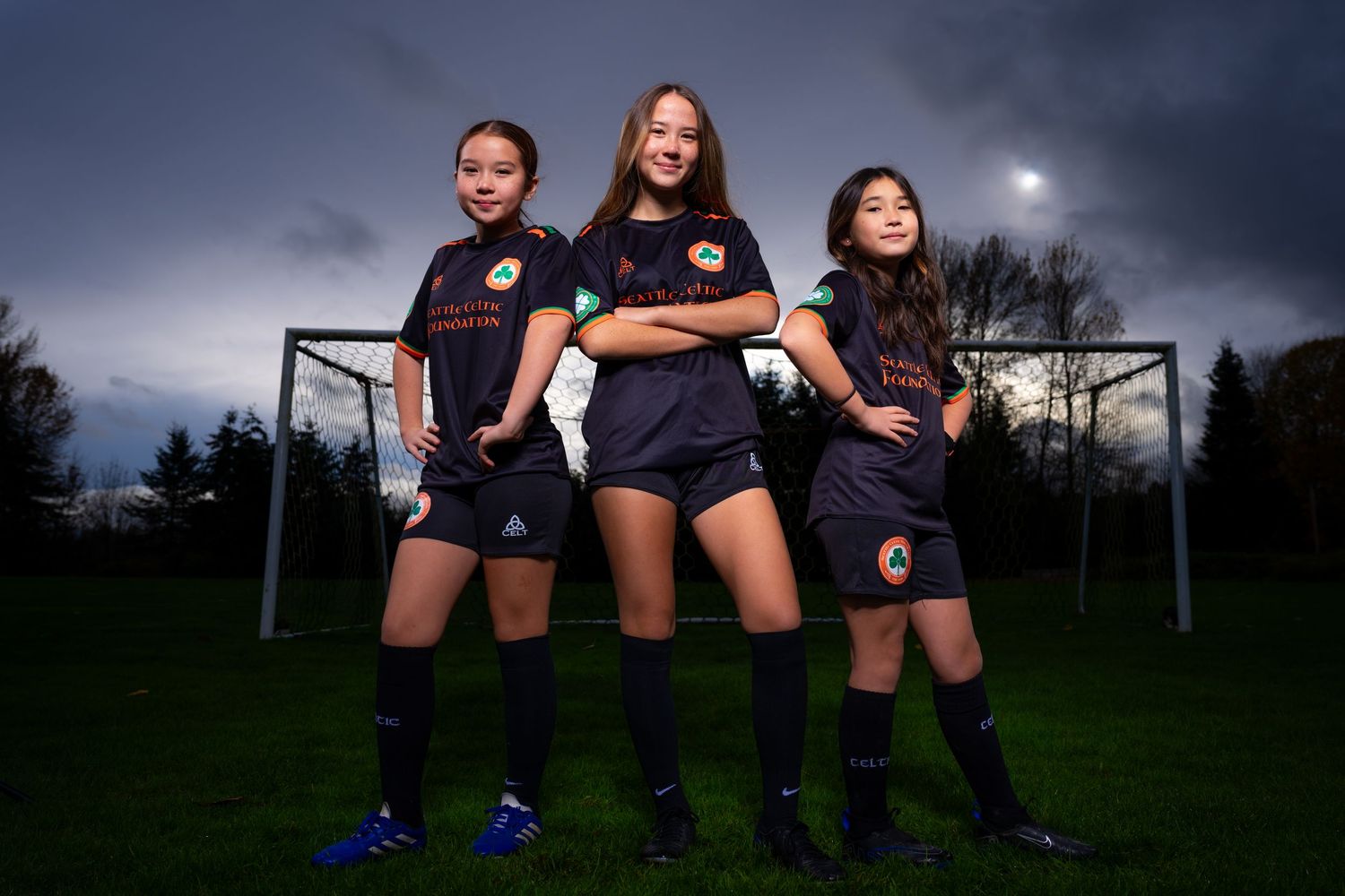 Three soccer players pose confidently in front of a goal on a dark moody evening with dramatic lighting.