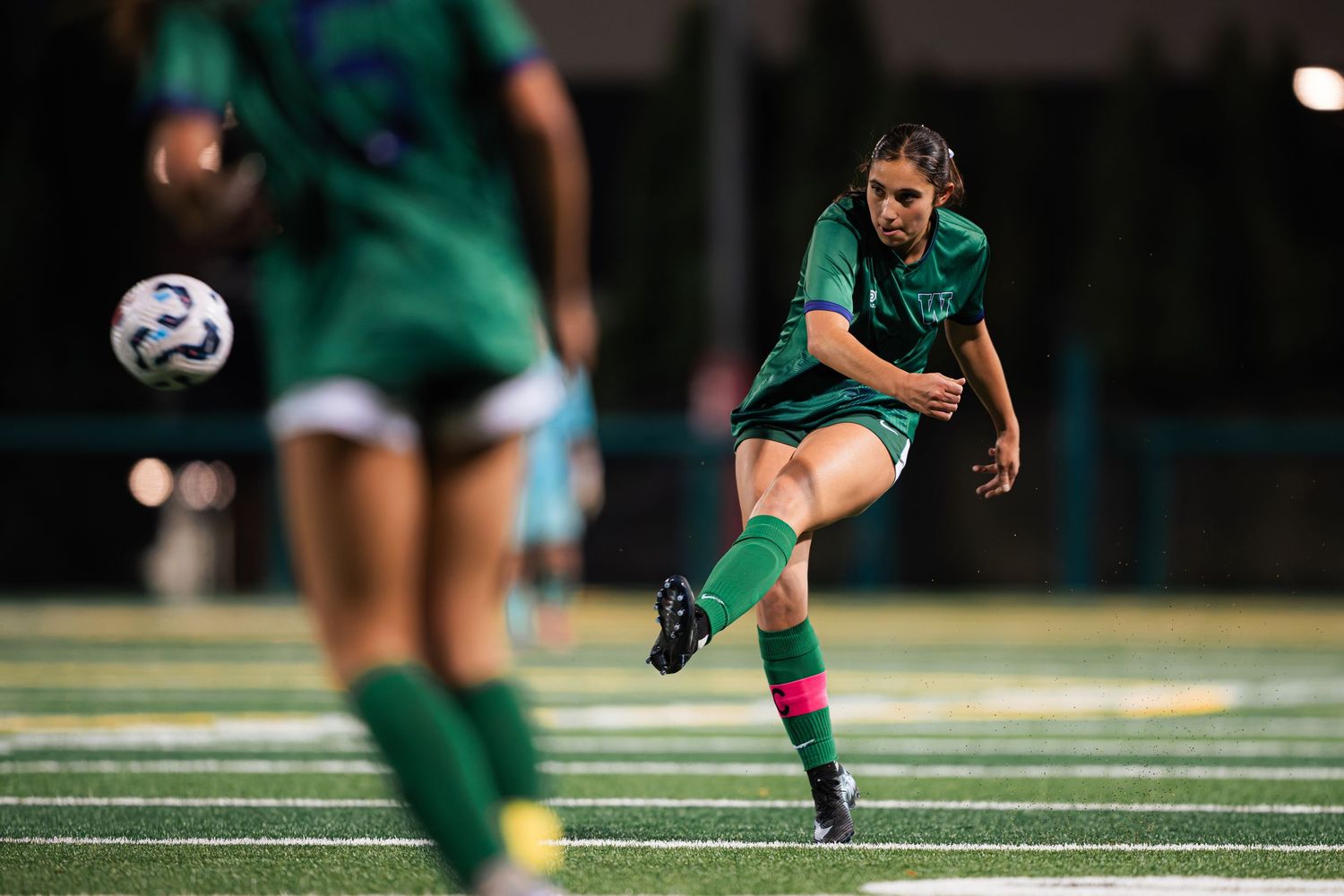 Soccer player in green uniform kicking the ball during a nighttime match on artificial turf field.