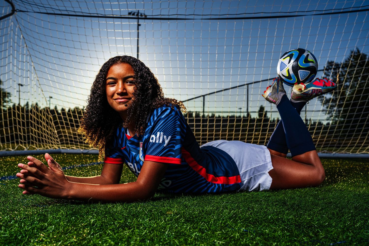 Soccer player lying in front of goal net on grass field during outdoor photoshoot.