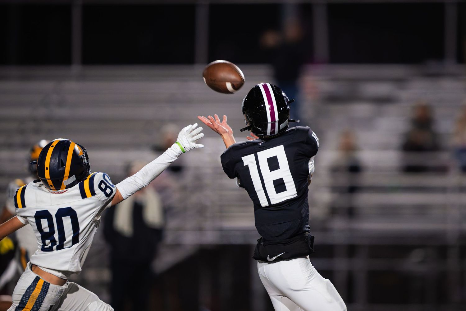 Two football players in action during a night game, with one player reaching to catch a pass against a defender.