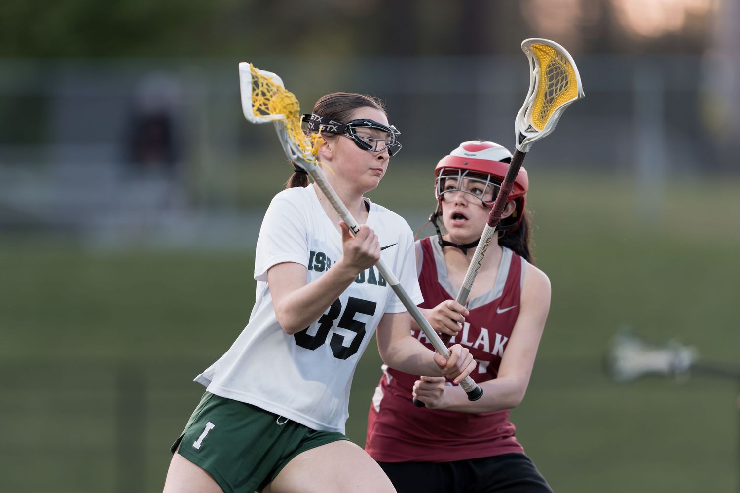 A lacrosse player in a white jersey makes a shot while being defended by a player in a maroon uniform.