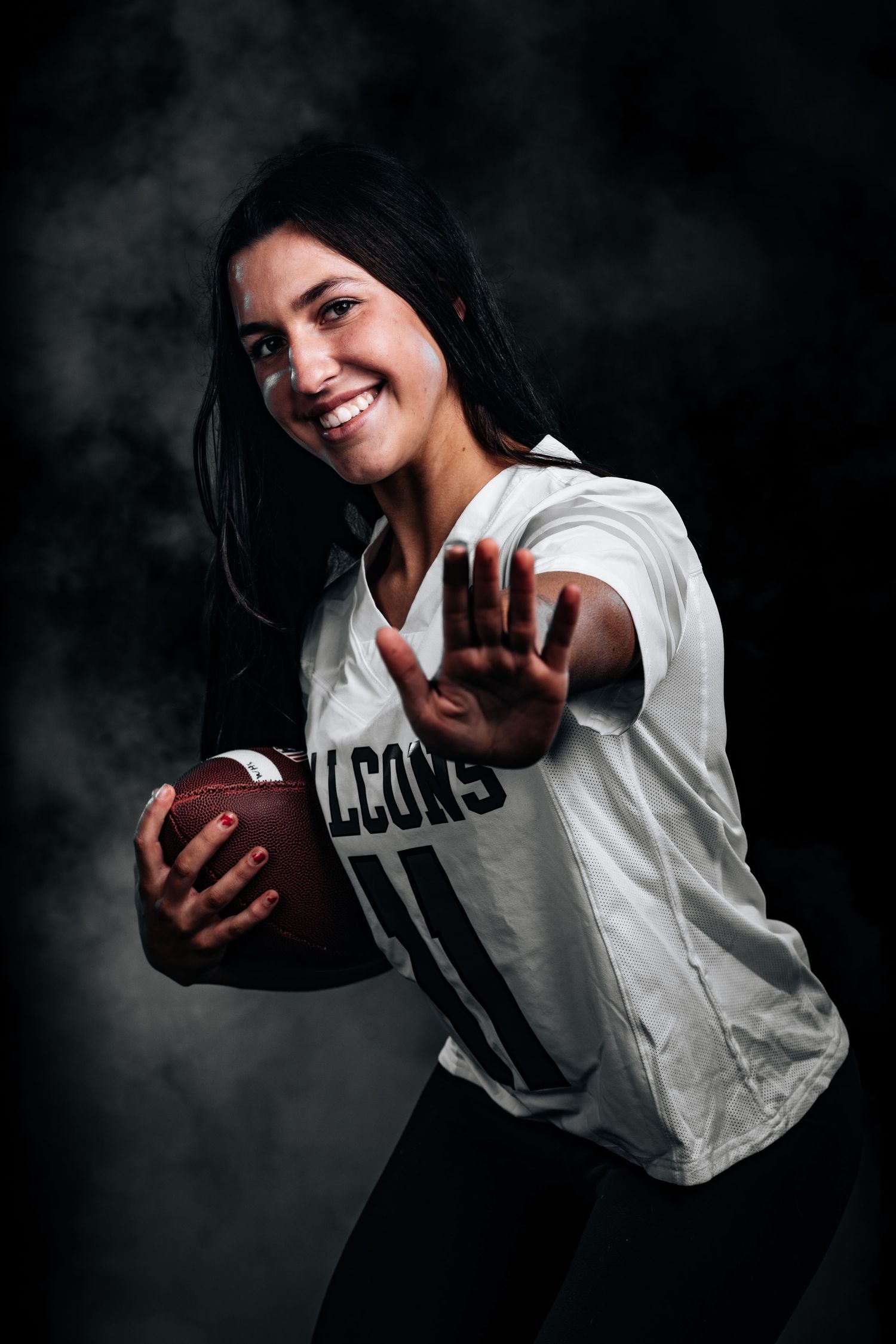 Athletic person in white jersey holds football and poses with outstretched hand against dark smoky background.