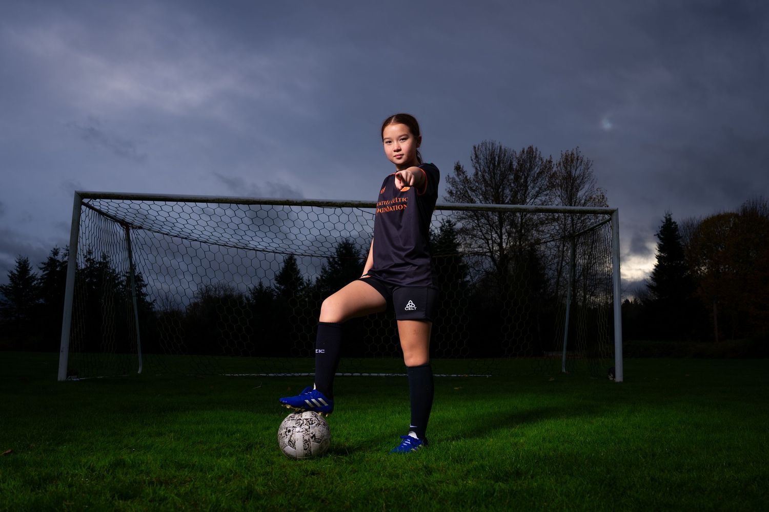 Soccer player poses with ball on field at dusk with goal post in background under dramatic sky.
